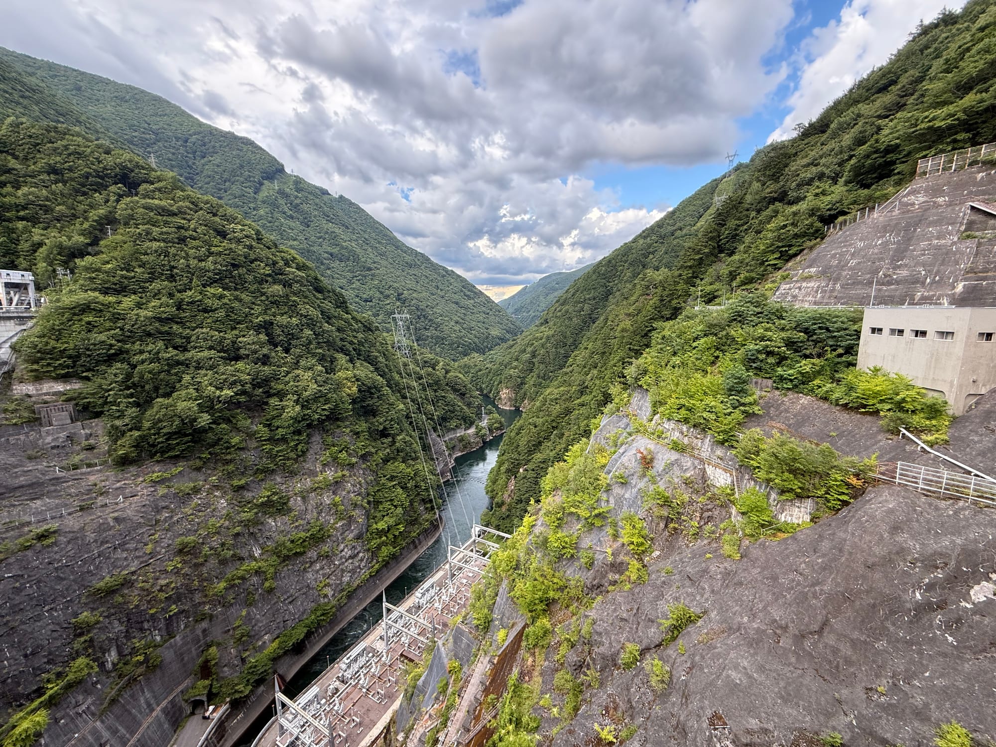 View downstream from Nagawado Dam, showing the narrow Azusa River gorge, hydroelectric facilities, steep forested mountainsides and dramatic clouds over the valley.