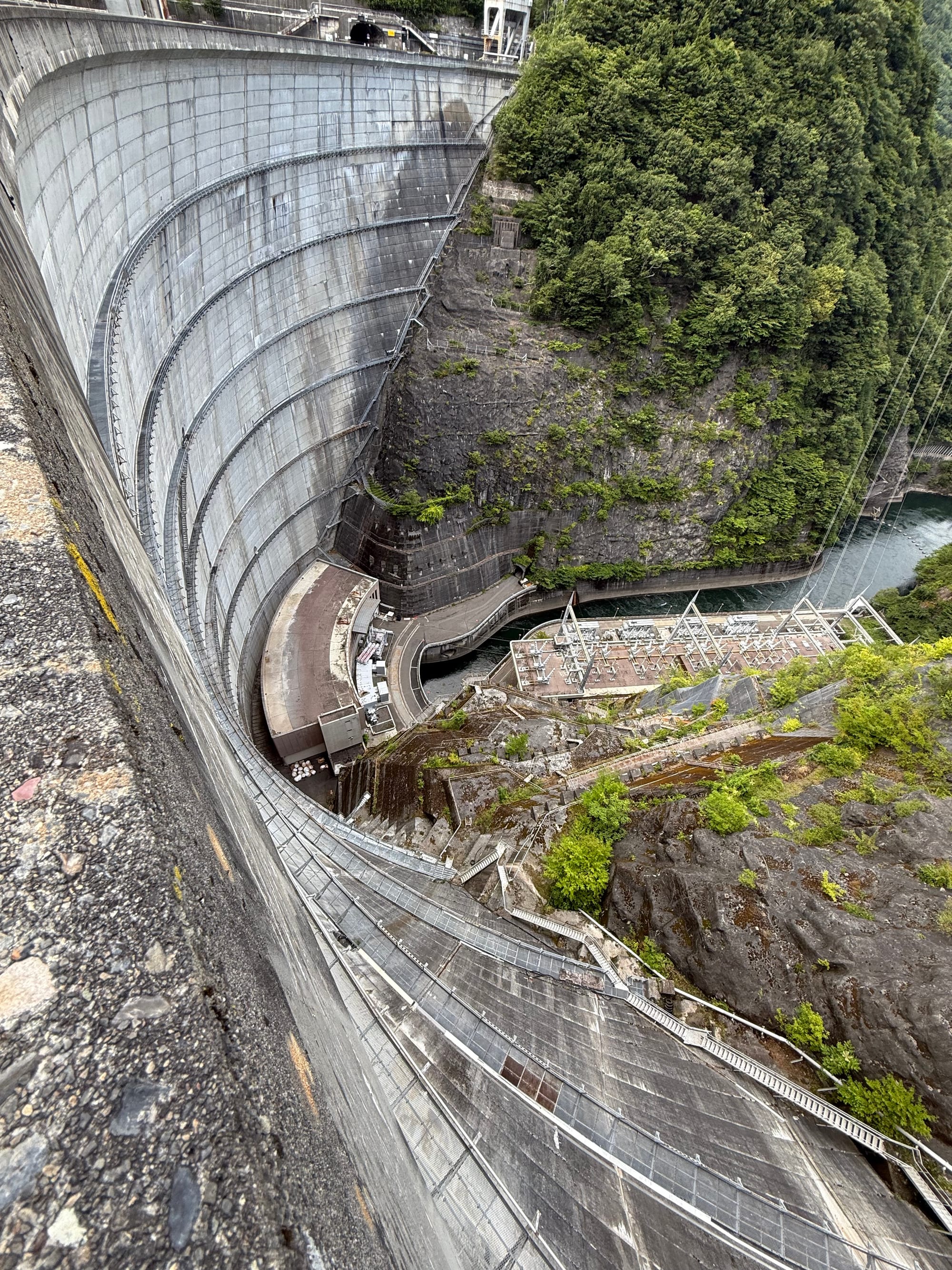 View down the curved concrete face of Nagawado Dam, showing the hydroelectric facilities below, the Azusa River gorge and steep forested mountainsides beyond.