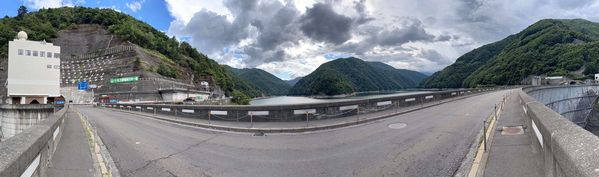 Panoramic view across Nagawado Dam, showing the reservoir, dam crest road, hydroelectric facilities and forested mountains beneath dramatic clouds in the Azusa River valley.