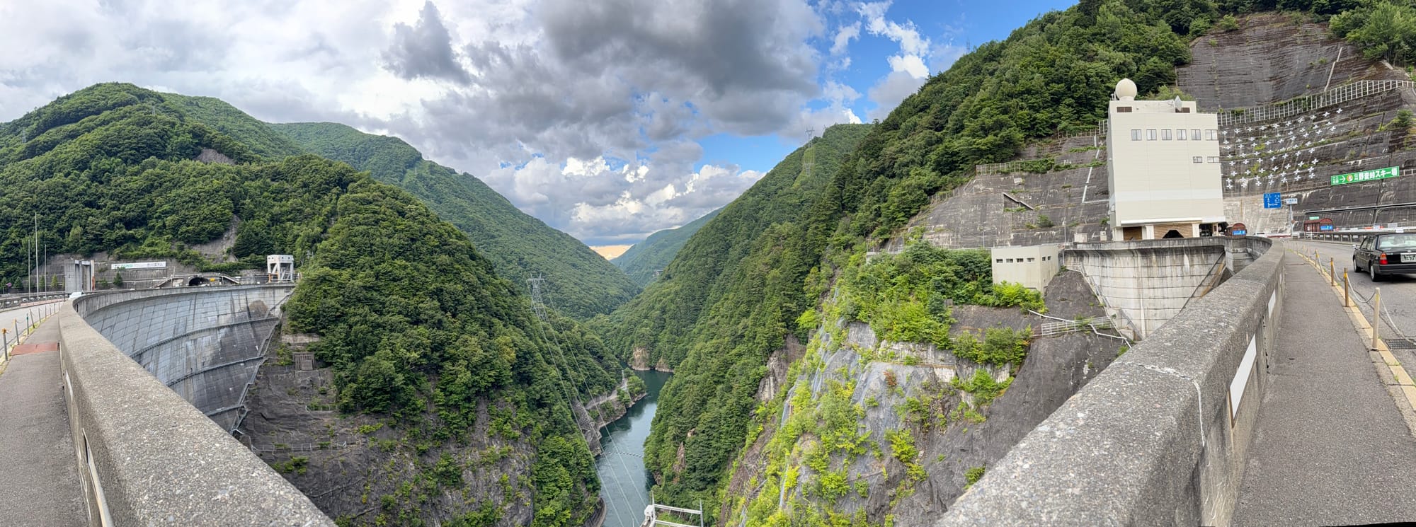 Panoramic view from Nagawado Dam looking downstream into the Azusa River gorge, with the curved dam wall, hydroelectric facilities and steep forested mountains on either side.