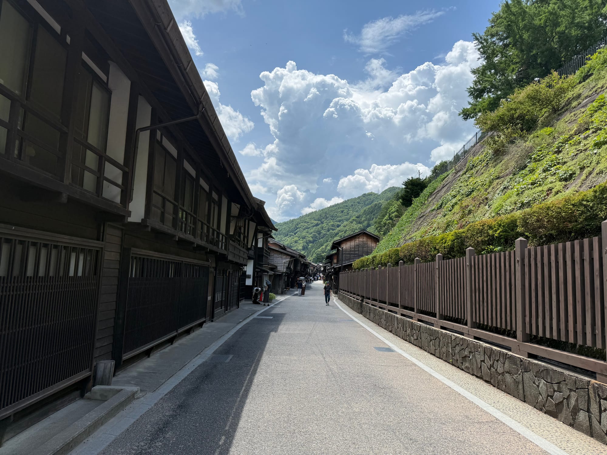 Traditional street in Narai-juku lined with preserved wooden buildings, with a mountain hillside rising beside the road beneath towering summer clouds and blue sky.