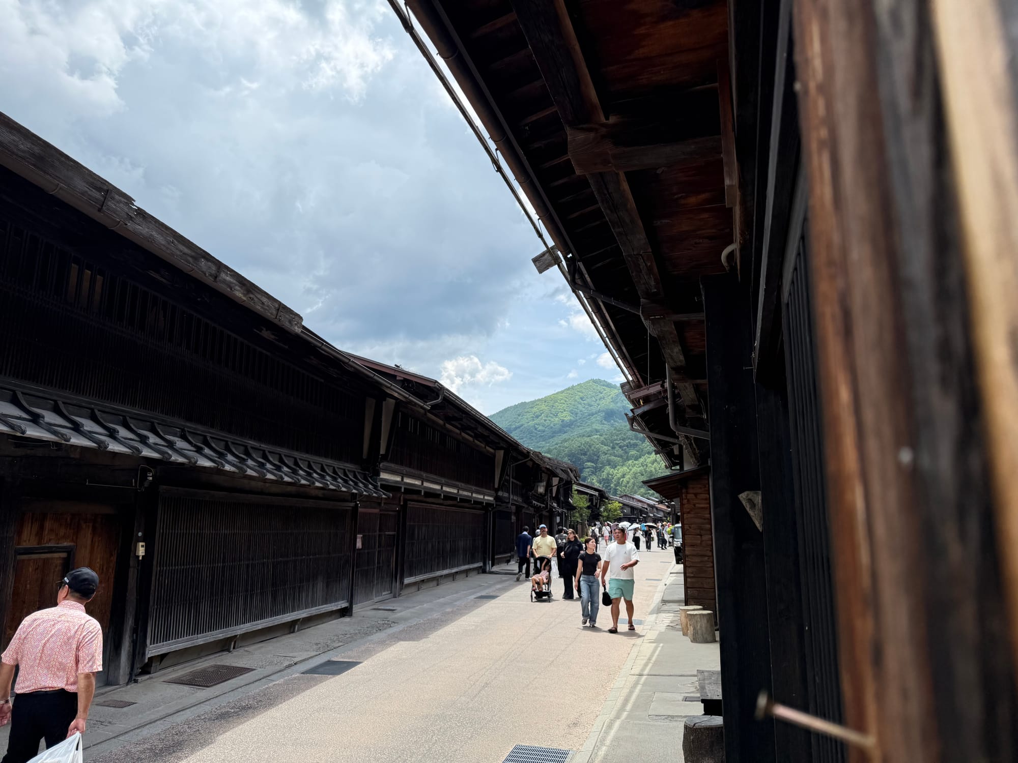 Visitors walk along the historic main street of Narai-juku, lined with preserved wooden merchant houses, with forested mountains rising beyond beneath a summer sky.