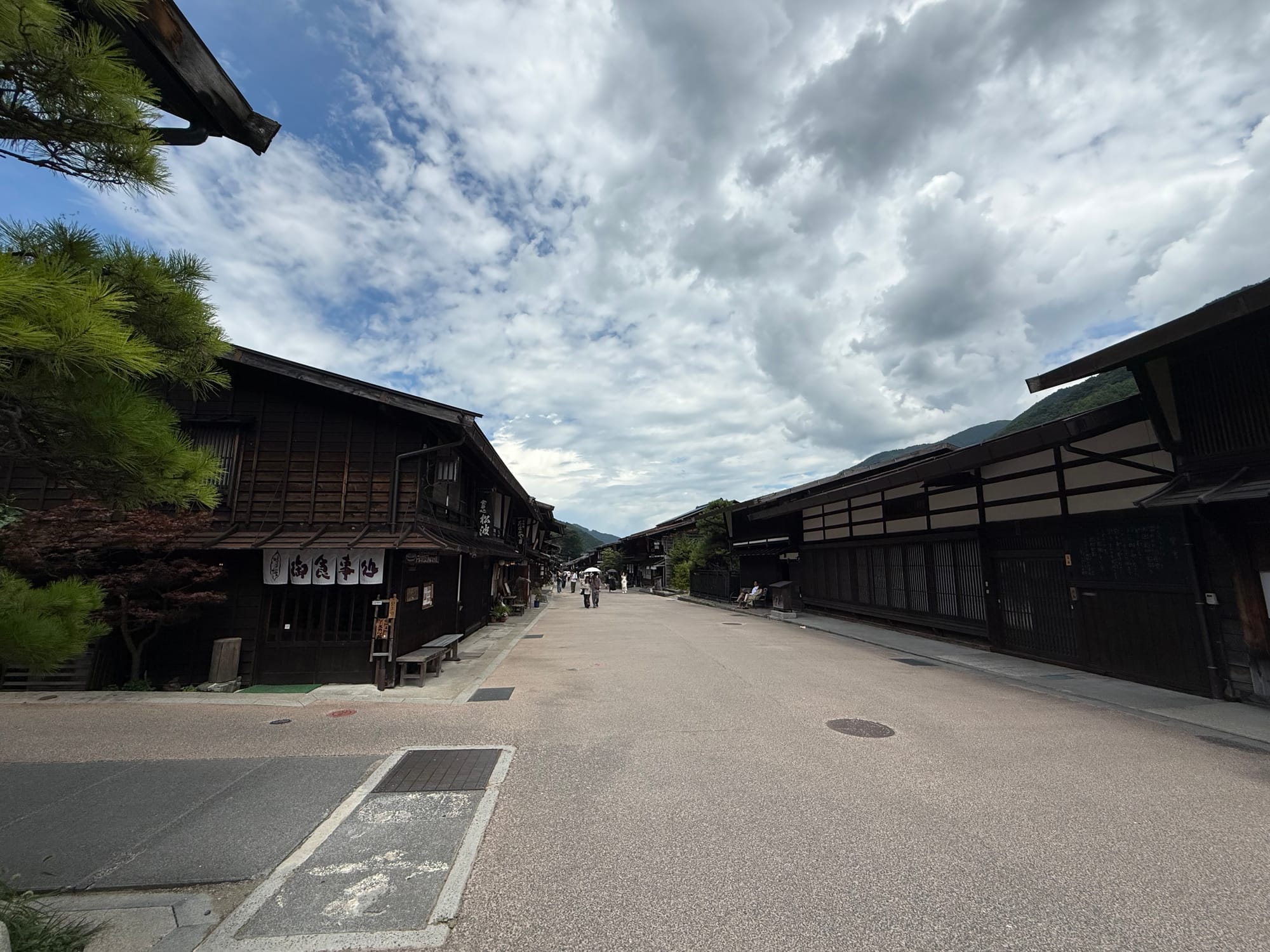 Wide view of Narai-juku's historic main street, lined with preserved wooden buildings and traditional shops beneath dramatic summer clouds and the surrounding mountains.
