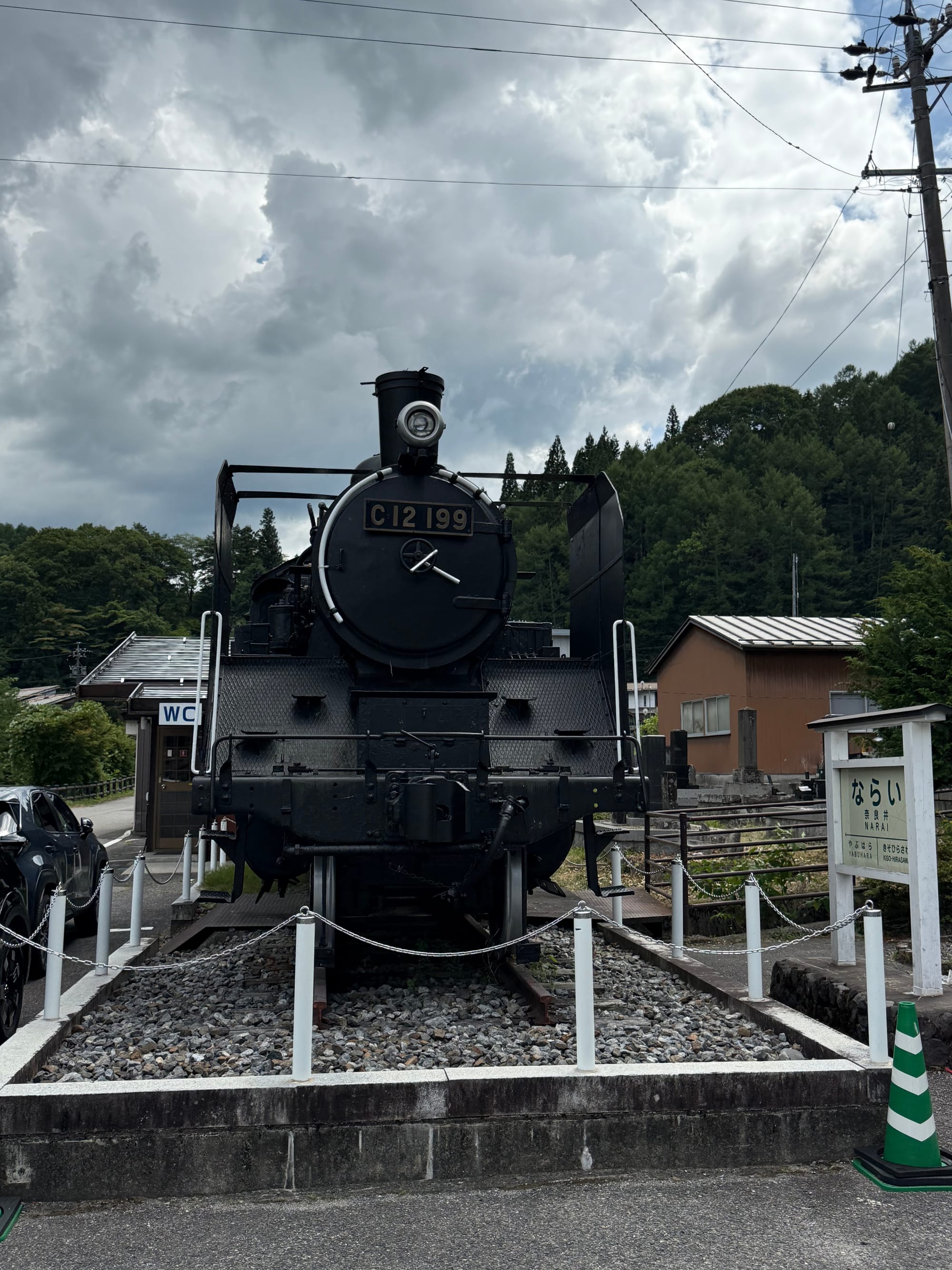 Preserved C12 199 steam locomotive displayed beside Narai Station, with the station sign, railway tracks and forested hills visible beneath dark summer clouds.