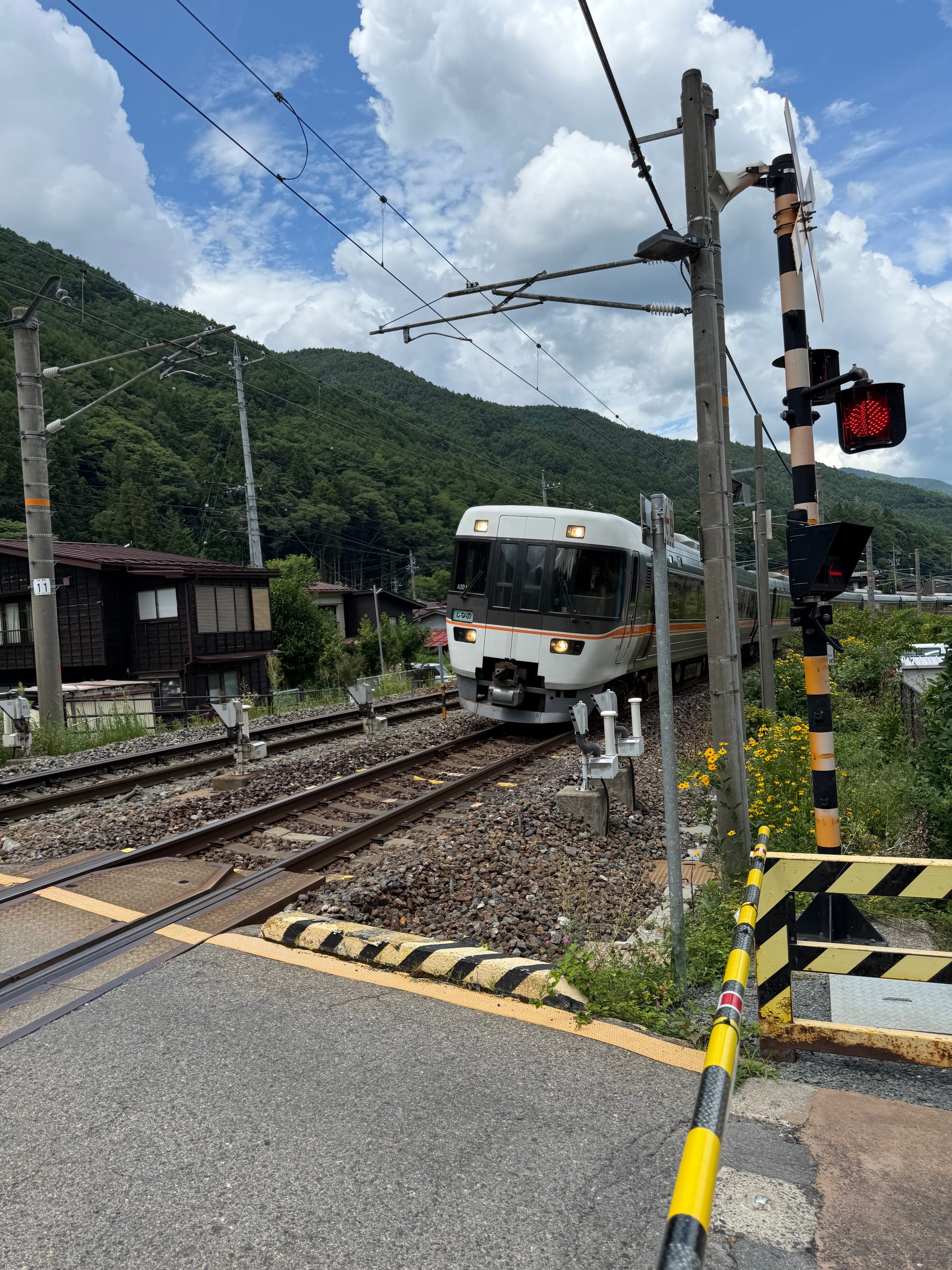 JR Central train crossing a level crossing near Narai Station, with forested mountains, overhead power lines and towering summer clouds rising above the Kiso Valley.