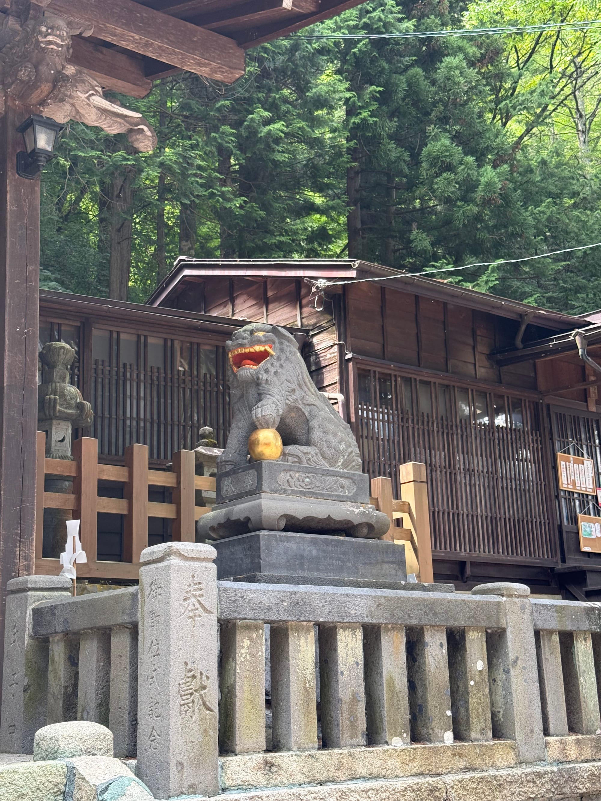 Stone komainu guardian statue at Shizume-Kinka Shrine, holding a golden sphere and standing before traditional wooden shrine buildings beneath towering cedar trees.