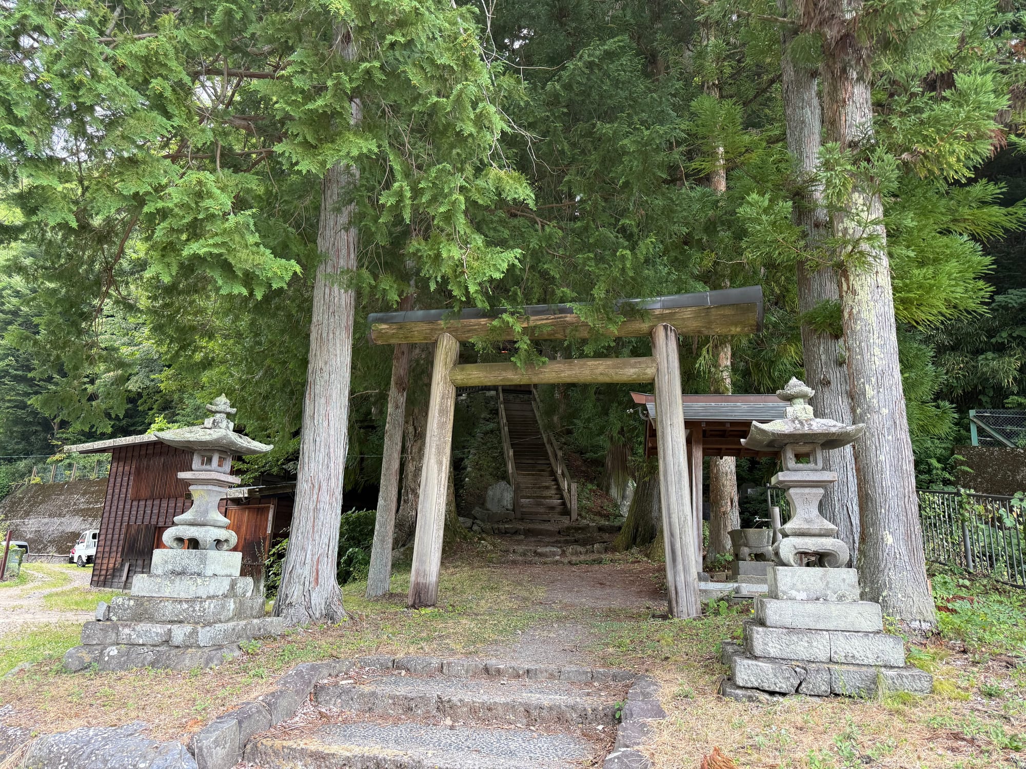 Wooden torii gate and stone lanterns marking the entrance to a small forest shrine near Narai-juku, with a steep staircase climbing through towering cedar trees.