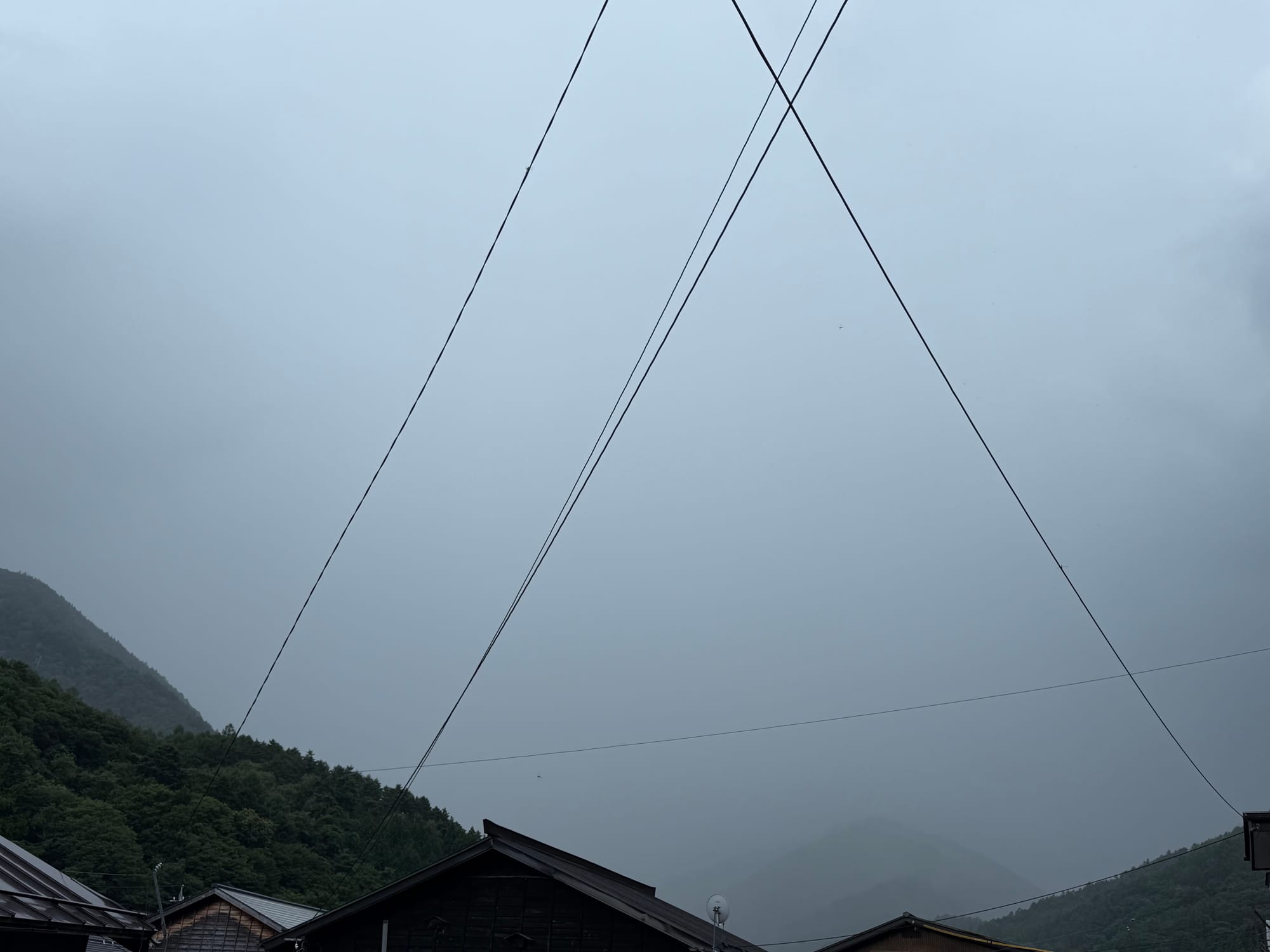 Low clouds and mist obscure the mountains around Narai-juku, with rooftops, forested slopes and overhead power lines visible beneath a grey rain-filled sky.