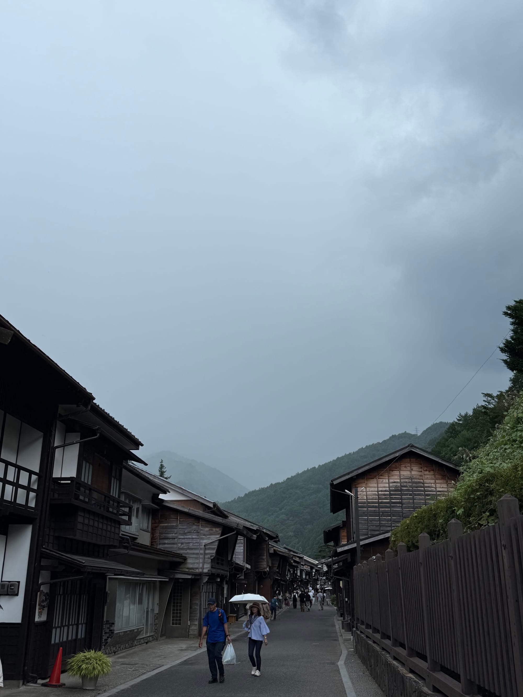 Historic street in Narai-juku under dark rain clouds, with preserved wooden buildings, visitors carrying umbrellas and mist-covered mountains rising beyond the village.