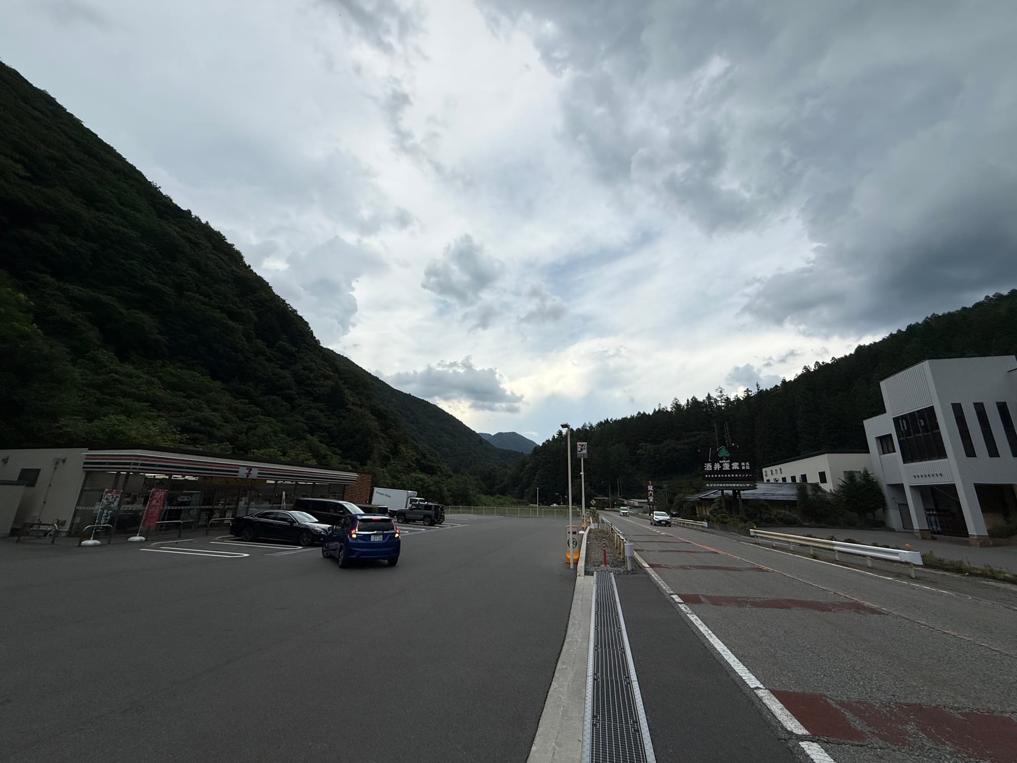 Mountain roadside service area in the Kiso Valley, with a convenience store, parked cars and forested slopes beneath dramatic storm clouds gathering over the surrounding peaks.