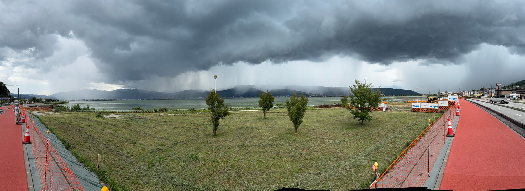 Panoramic view across Lake Suwa beneath dramatic storm clouds, with rain shafts falling over distant mountains, lakeside promenades and open green space in the foreground.
