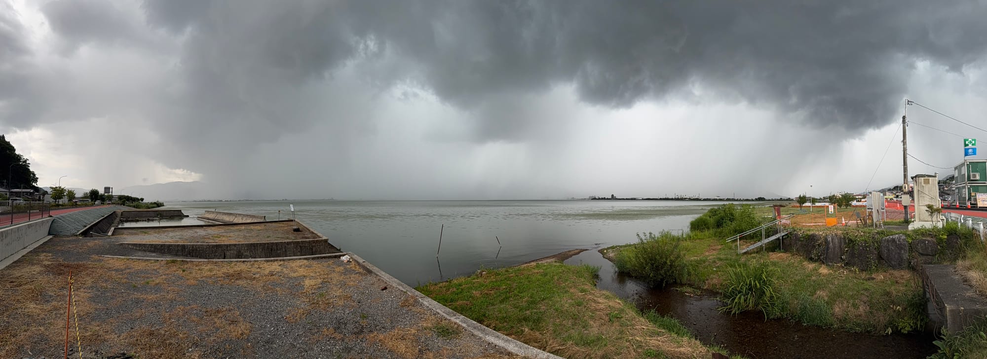 Panoramic view across Lake Suwa beneath a severe summer storm, with dark cloud bases, heavy rain shafts over the lake, distant mountains and a small stream entering the shoreline.