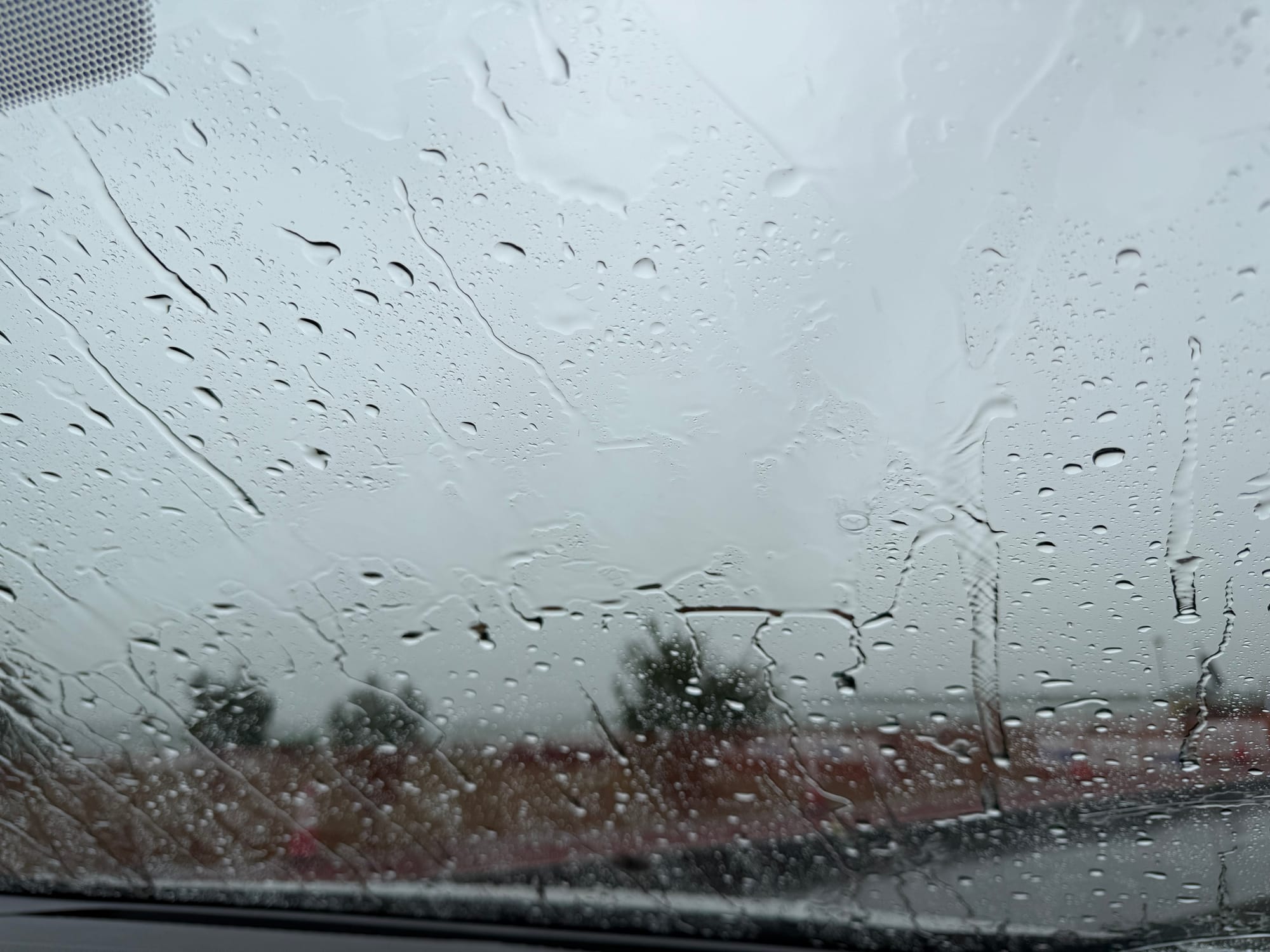 Raindrops streaming down a car window during a thunderstorm at Lake Suwa, obscuring the lakeshore and distant landscape behind a curtain of heavy summer rain.