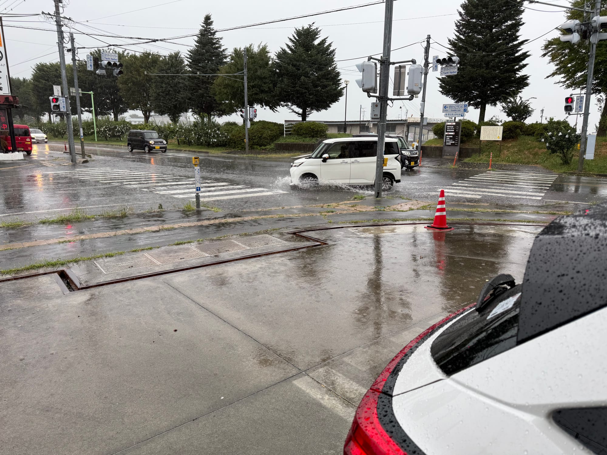 Heavy rain at a lakeside intersection near Lake Suwa, with vehicles splashing through pooled water while viewed from outside a laundrette during a summer storm.
