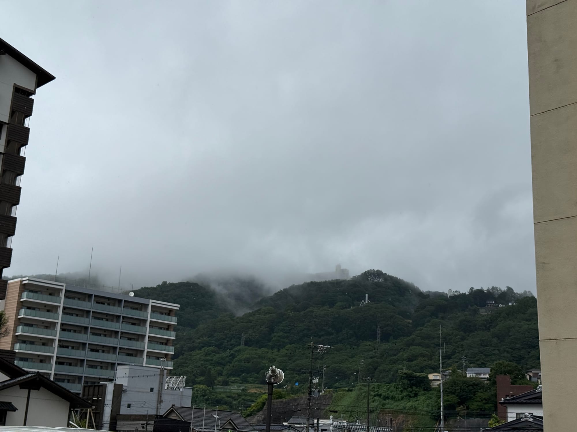 View from a hotel room in Suwa, showing low cloud and mist draped across forested hills after a summer storm, with nearby buildings partially framed by the window view.