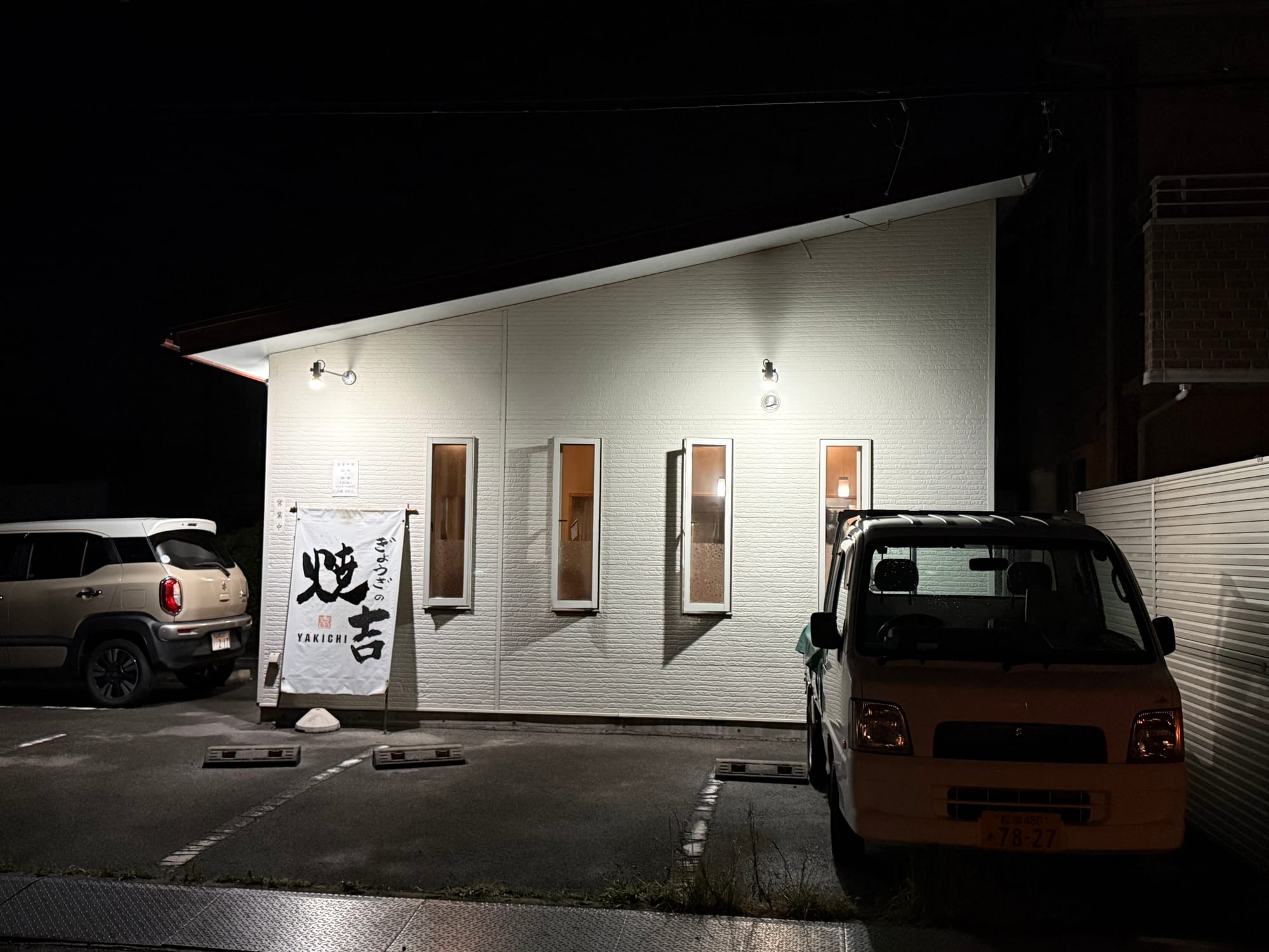 Yakichi Gyoza restaurant at night in Suwa, featuring a brightly lit white building, parked vehicles and the restaurant's banner sign outside the entrance.