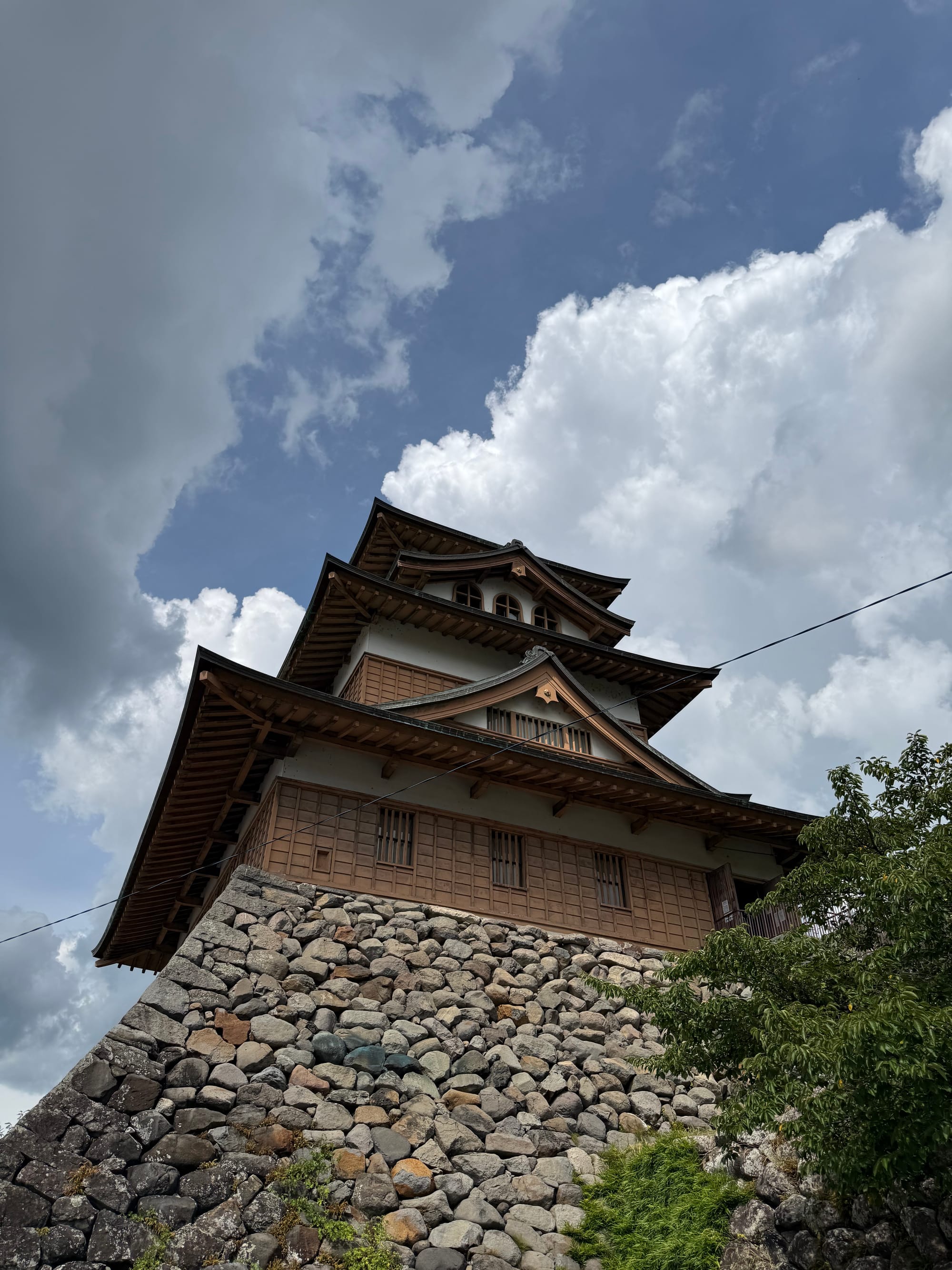 Takashima Castle keep in Suwa, viewed from below, showing its wooden upper levels and stone foundations beneath towering summer clouds and patches of blue sky.