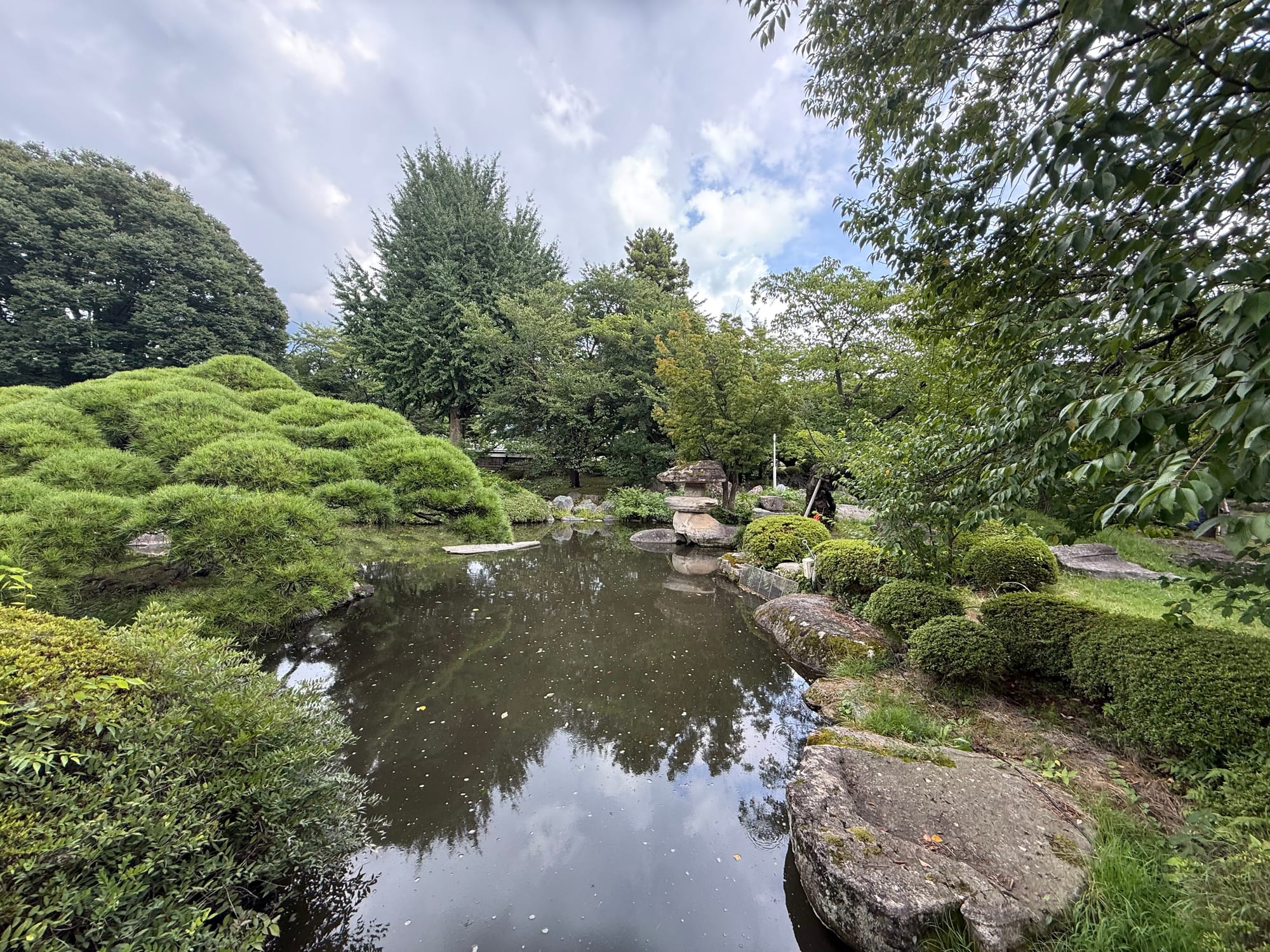 Japanese garden within Takashima Castle Park, featuring a reflective pond, carefully pruned pine trees, stone lanterns, trimmed shrubs and mature trees beneath a cloudy sky.