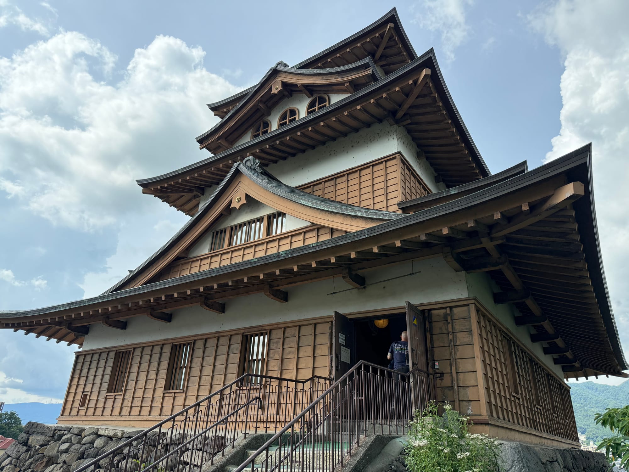 Takashima Castle keep in Suwa, viewed from the entrance staircase, featuring traditional wooden architecture, sweeping tiled roofs and a dramatic summer sky overhead.
