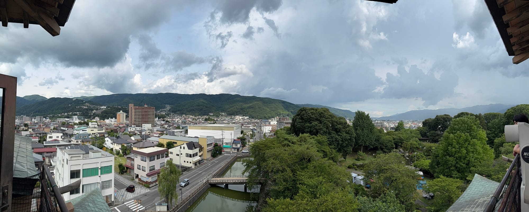 Panoramic view from Takashima Castle overlooking Suwa, with the castle park, canal, town streets and surrounding mountains beneath dramatic summer storm clouds.