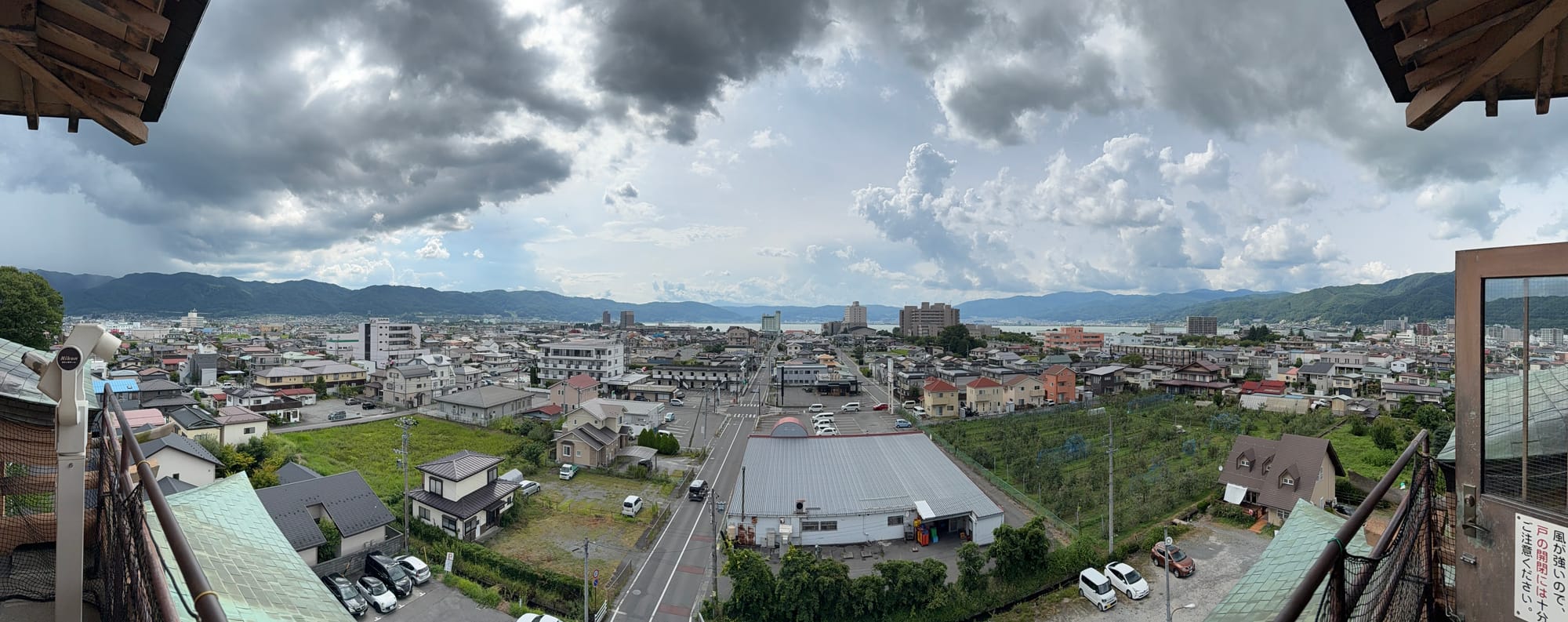 Panoramic view from Takashima Castle overlooking Suwa and Lake Suwa, with residential neighbourhoods, farmland, surrounding mountains and dramatic storm clouds gathering overhead.