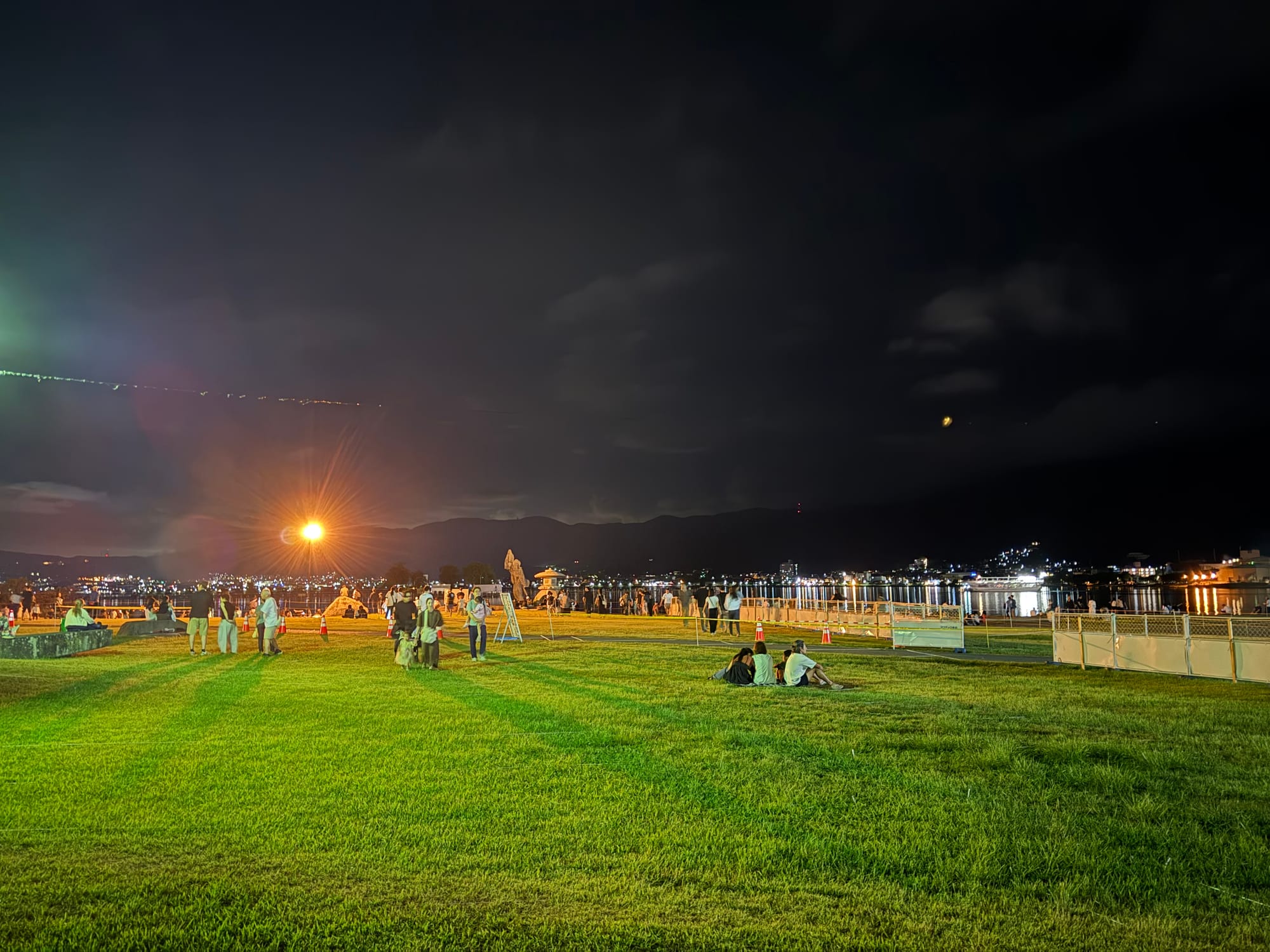 Visitors gather on the lakeside lawn at Lake Suwa after dark, with town lights reflecting on the water and mountains silhouetted in the distance before a fireworks display.