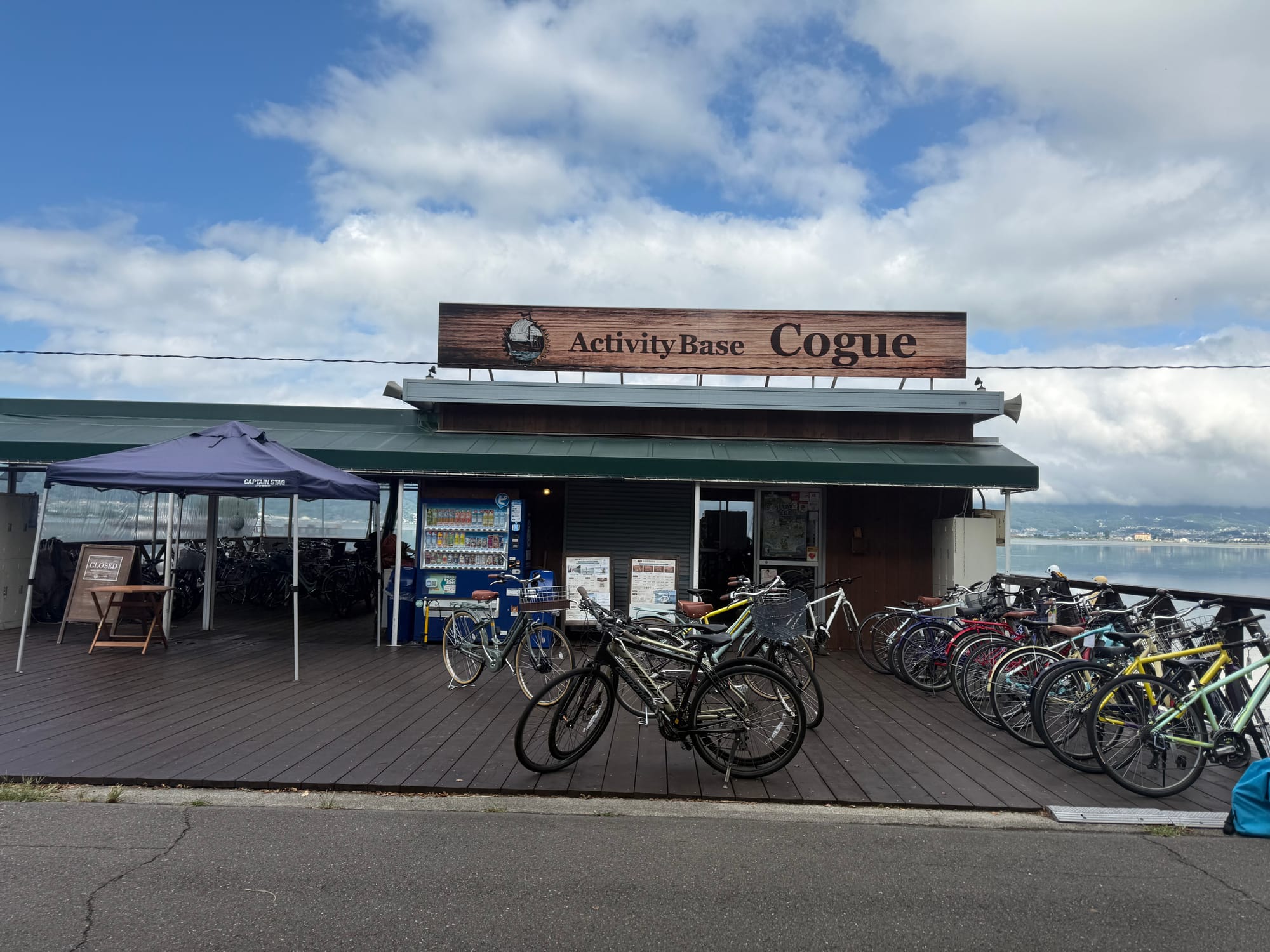 Activity Base Cogue on the shore of Lake Suwa, featuring bicycle rentals, outdoor decking, vending machines and rows of parked bicycles beneath a partly cloudy sky.