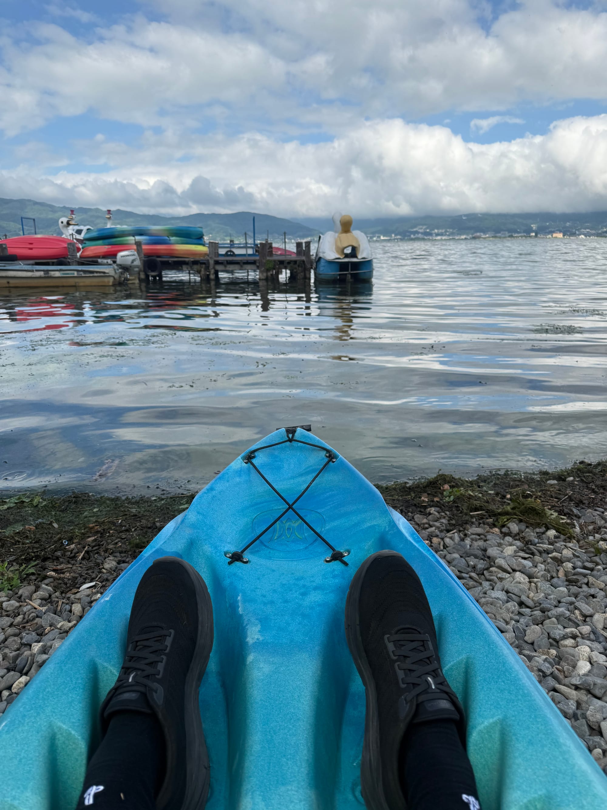 Kayak pulled up on the shore of Lake Suwa before launch, with calm water, a lakeside jetty, stacked kayaks and mountains beneath a sky filled with towering clouds.