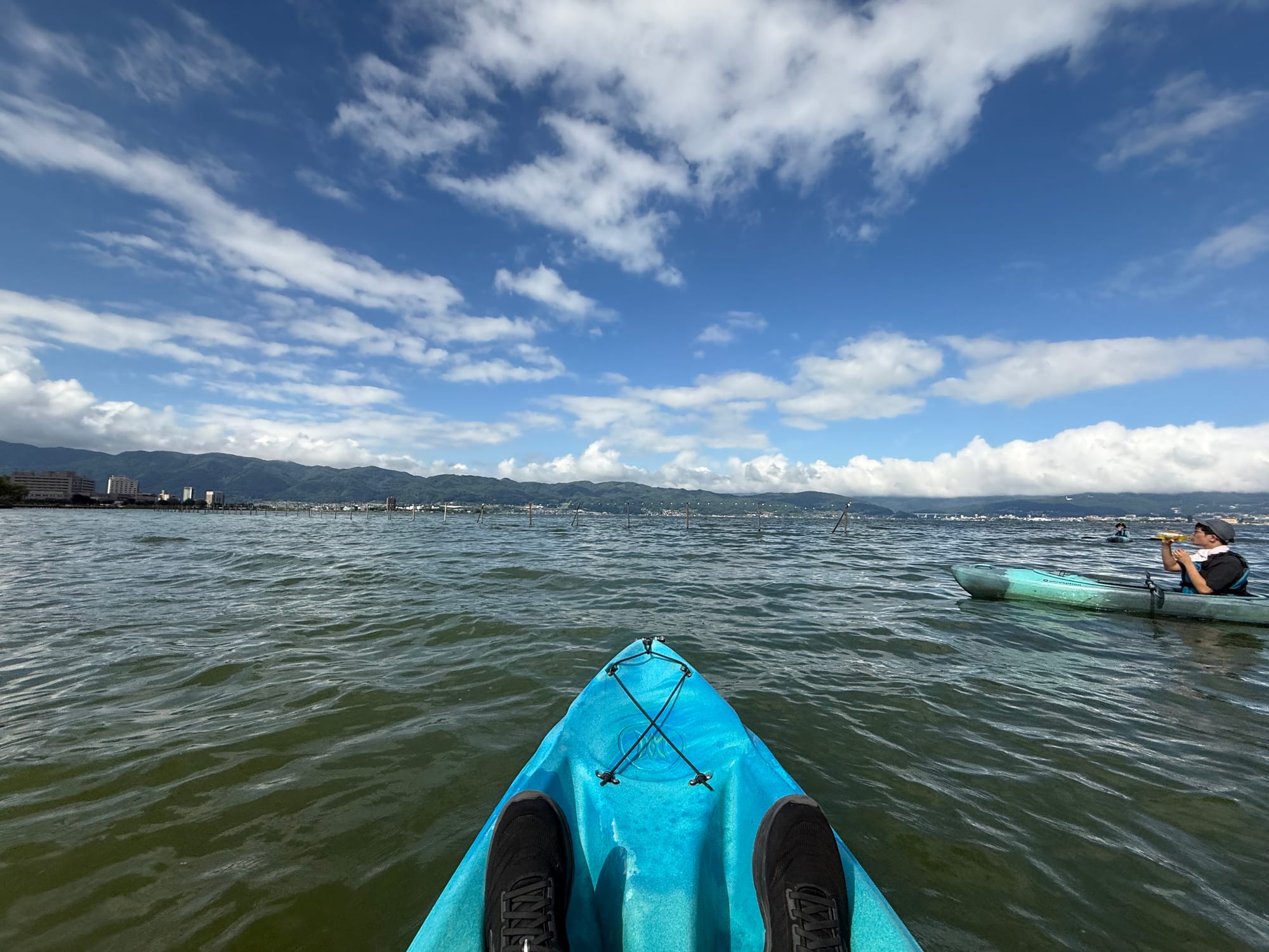 View from a kayak on Lake Suwa, with rippling water, surrounding mountains, another kayaker and dramatic clouds stretching across the sky above the lake.