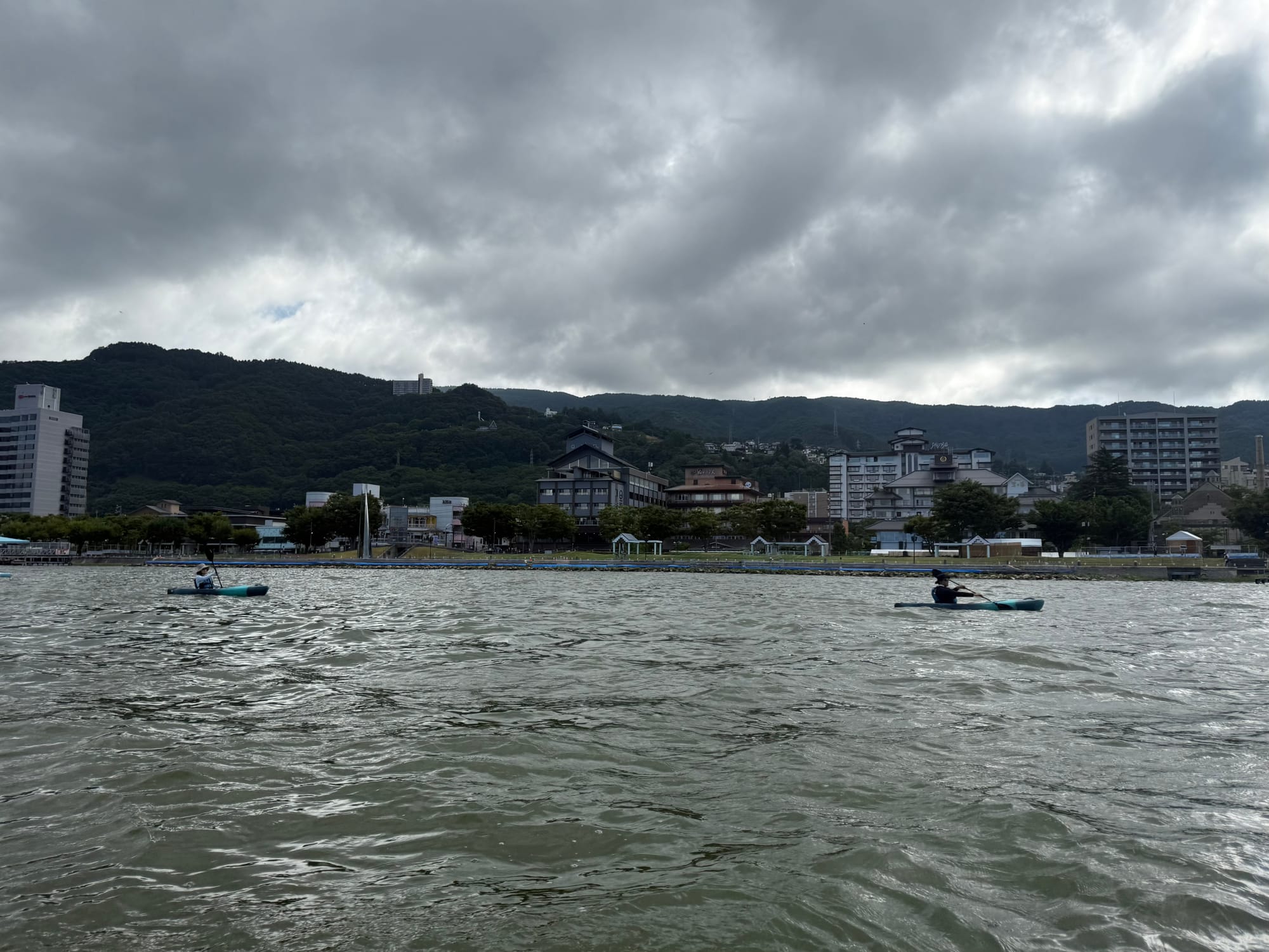 Kayakers on Lake Suwa beneath dark storm clouds, with the lakeside waterfront, hotels and forested hills of the Suwa Basin visible across the water.