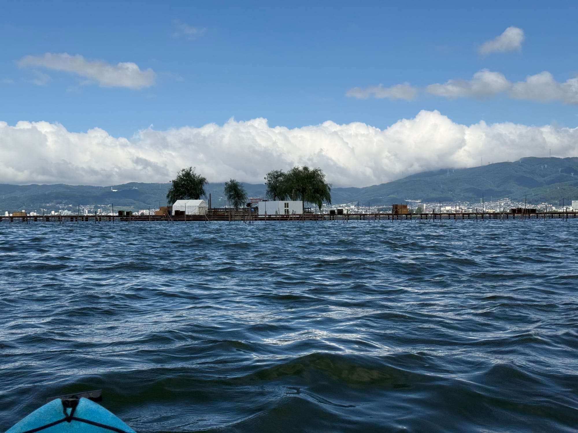 Copilot said:  Alt text (191 characters or under):  Fireworks launch platform on Lake Suwa viewed from a kayak, with staging structures, calm lakeside facilities and mountains rising beneath towering summer clouds.