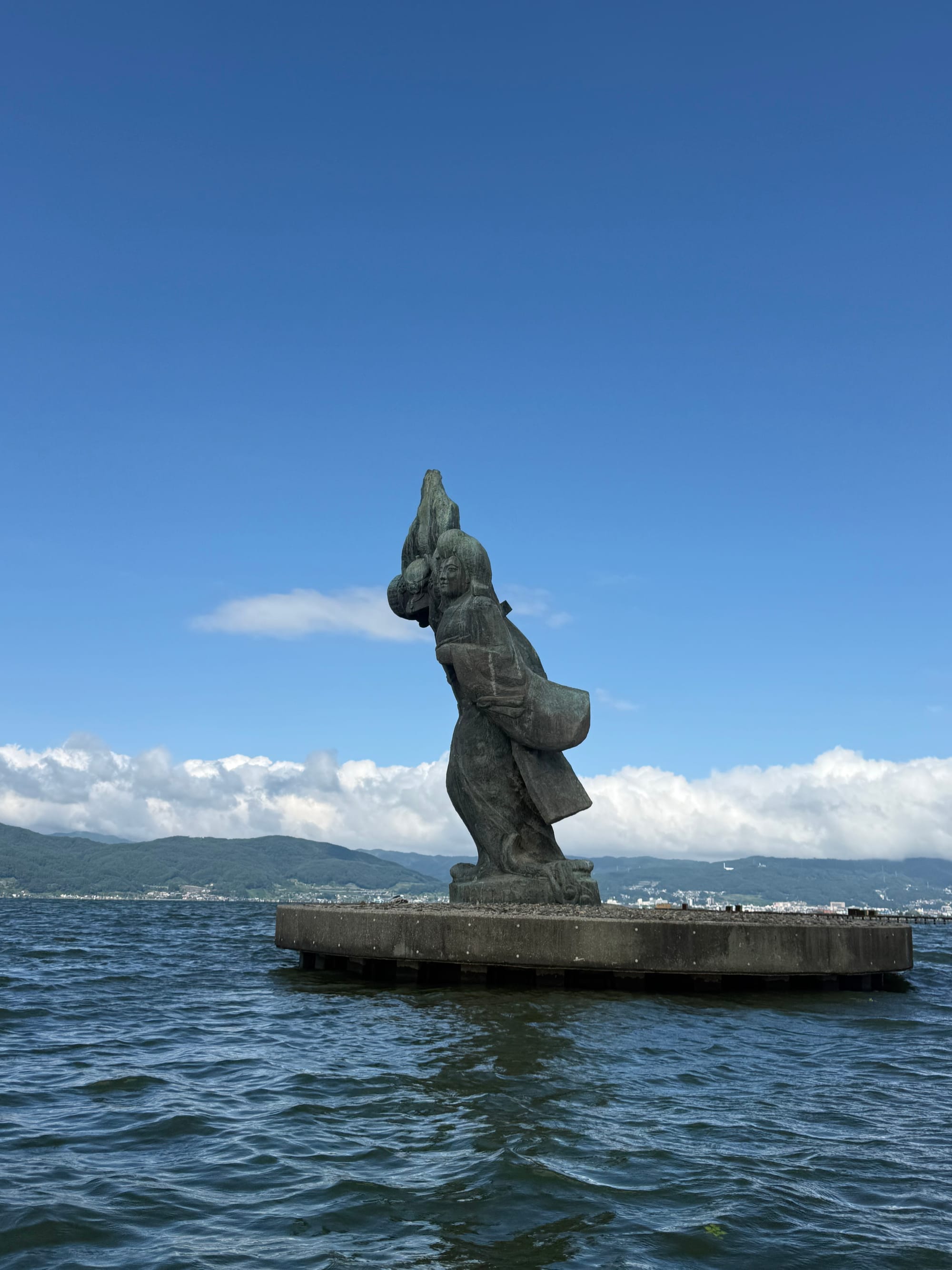 Statue of the Maiden of Lake Suwa standing on a platform in Lake Suwa, with rippling water, distant mountains and towering clouds beneath a bright summer sky.