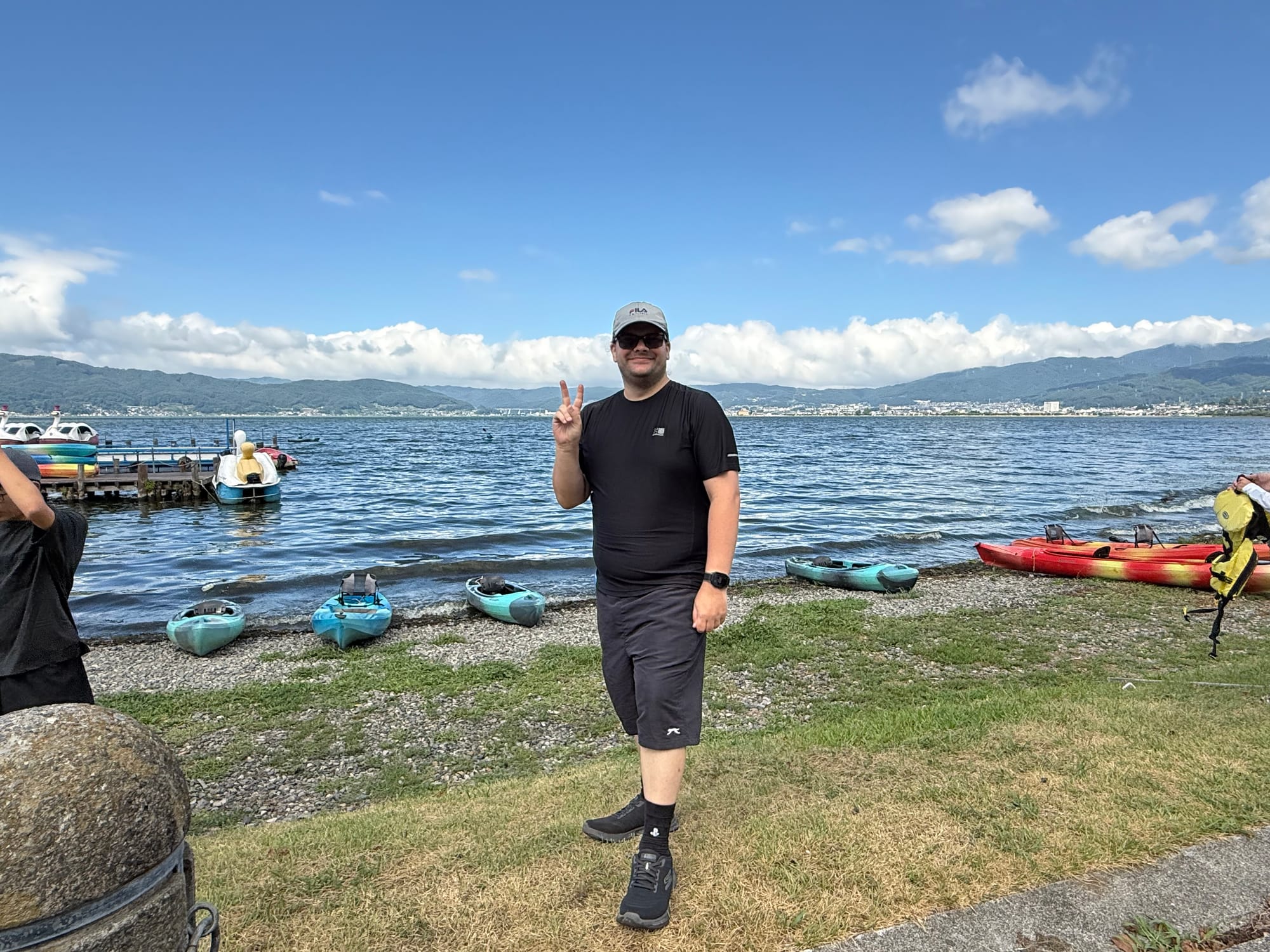 Visitor standing beside Lake Suwa after a kayaking session, with colourful kayaks on the shore, not so calm water and mountains stretching across the far side of the lake.