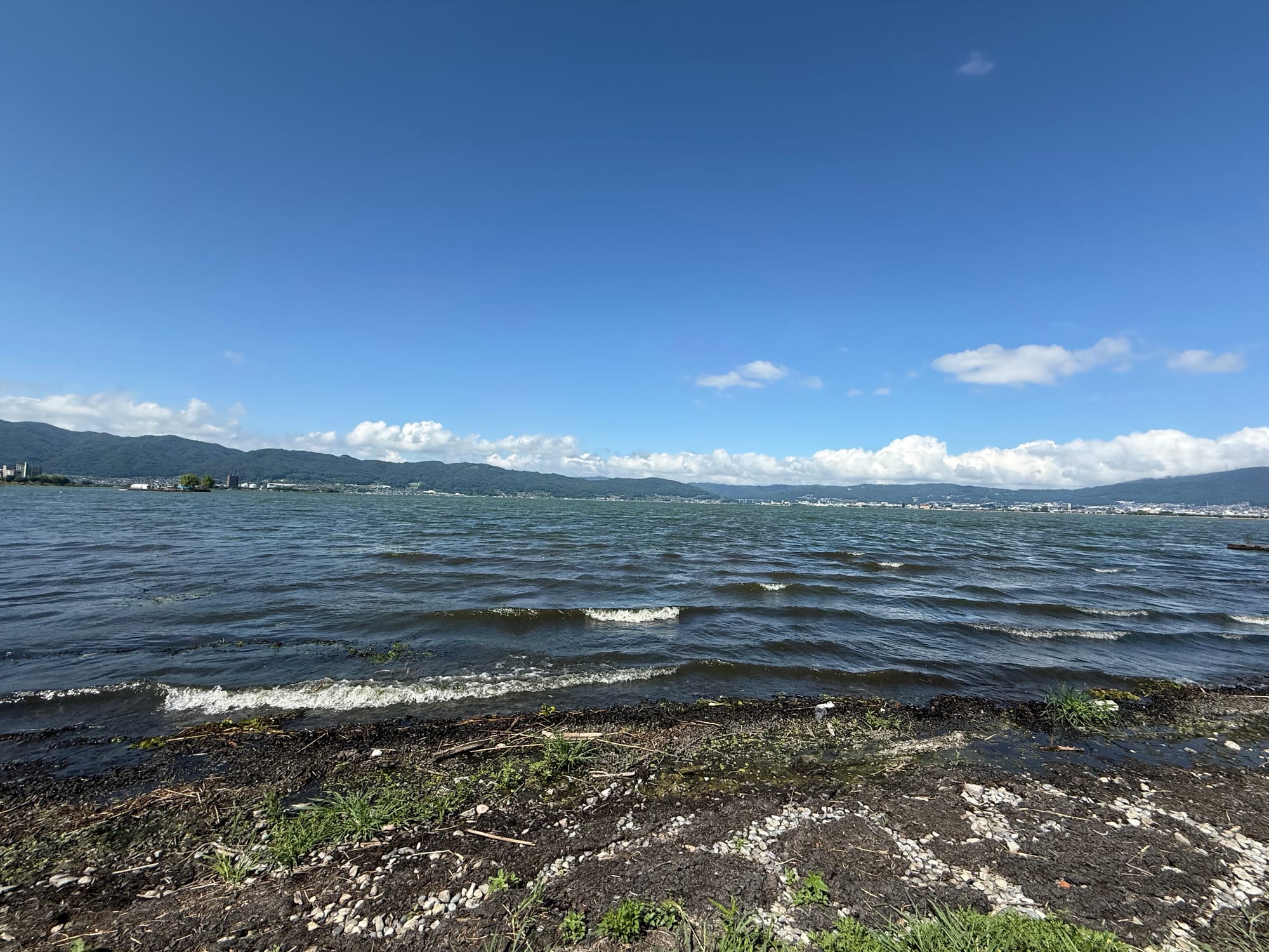 View across Lake Suwa from the shoreline, with small waves breaking along the pebbled shore, distant lakeside towns and mountains beneath a bright summer sky.