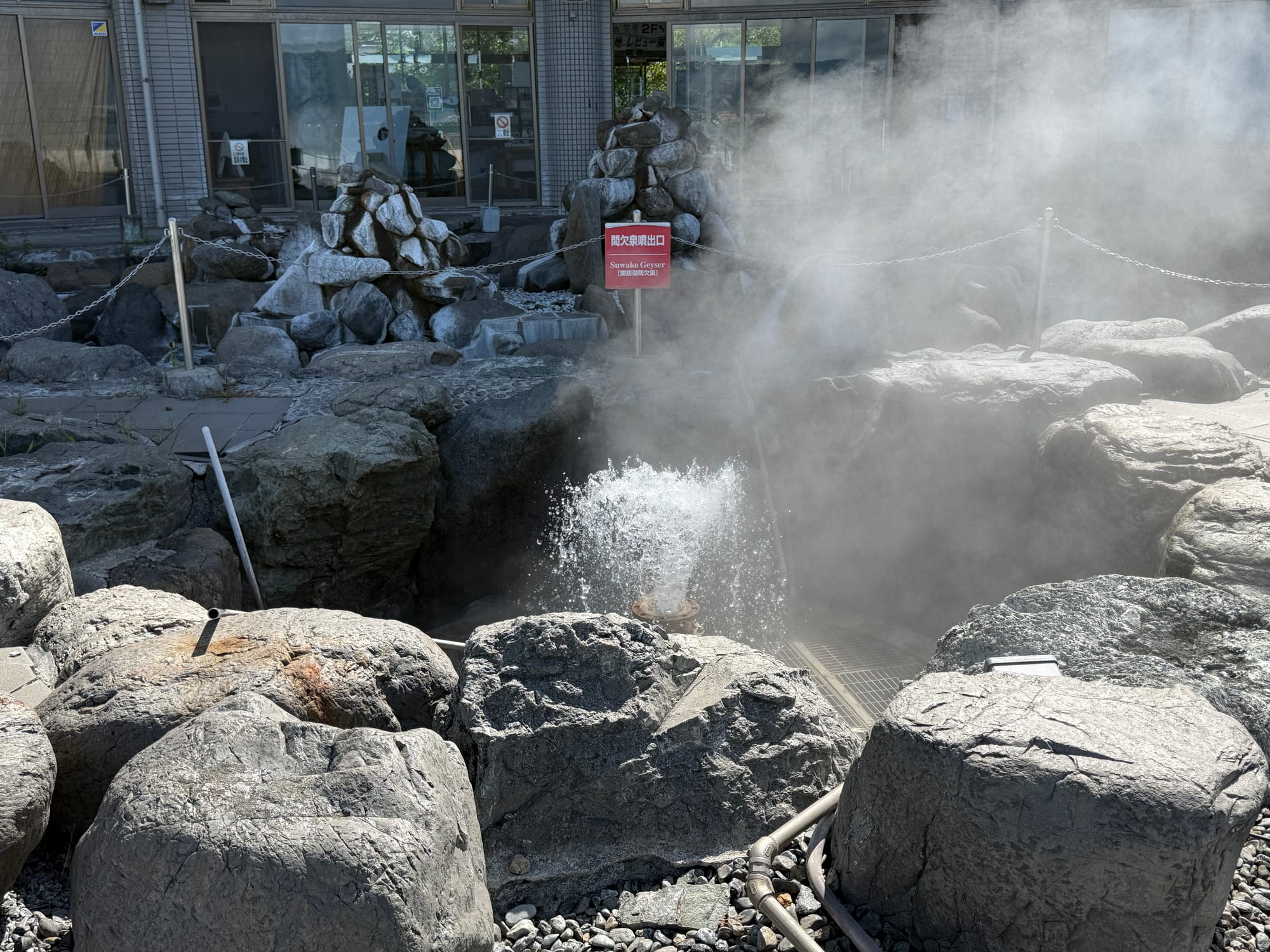 Suwa Geyser hot spring outlet near Lake Suwa, with steam rising from boiling water erupting among large rocks, surrounded by buildings and geothermal facilities.