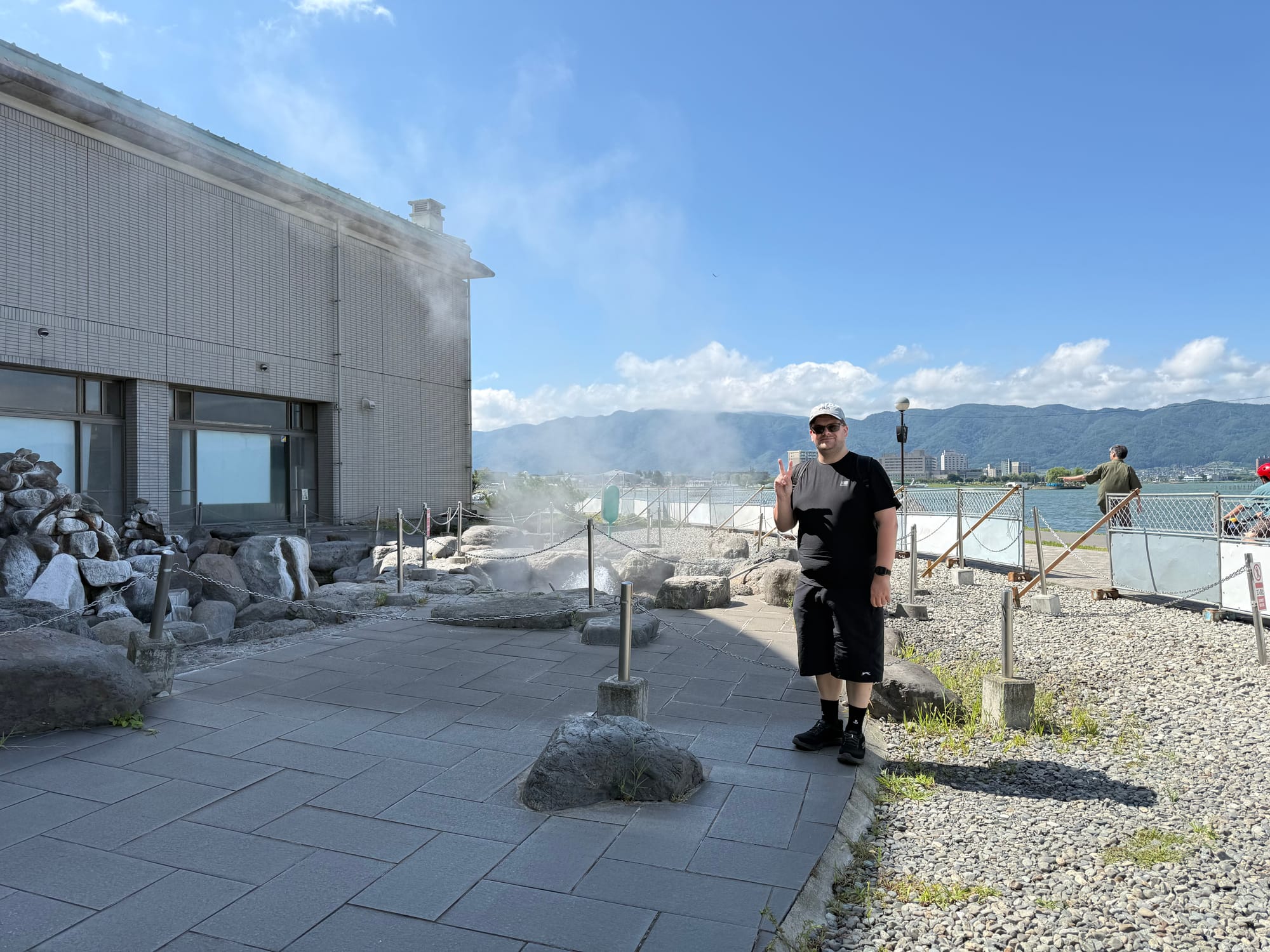 Visitor standing beside the steaming Suwa Geyser hot spring outlet near Lake Suwa, with geothermal vents, large rocks, lakeside scenery and mountains in the background.