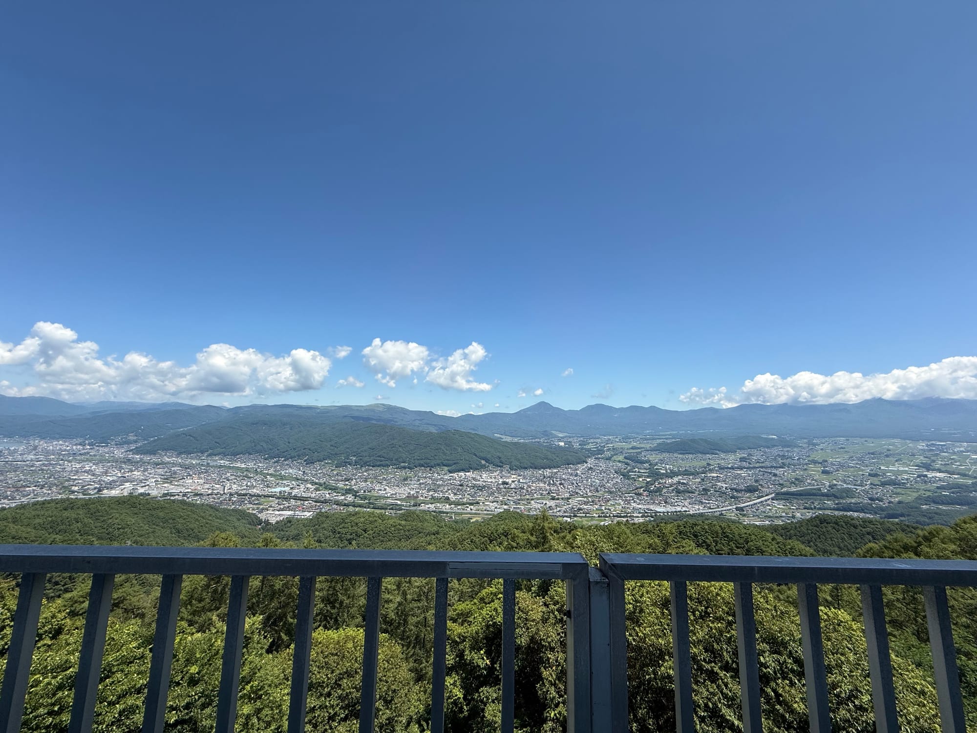anoramic view from the Tsuetsukitoge Observation Deck restaurant, overlooking the Suwa Basin, surrounding mountains and towns beneath a clear blue sky with scattered clouds.