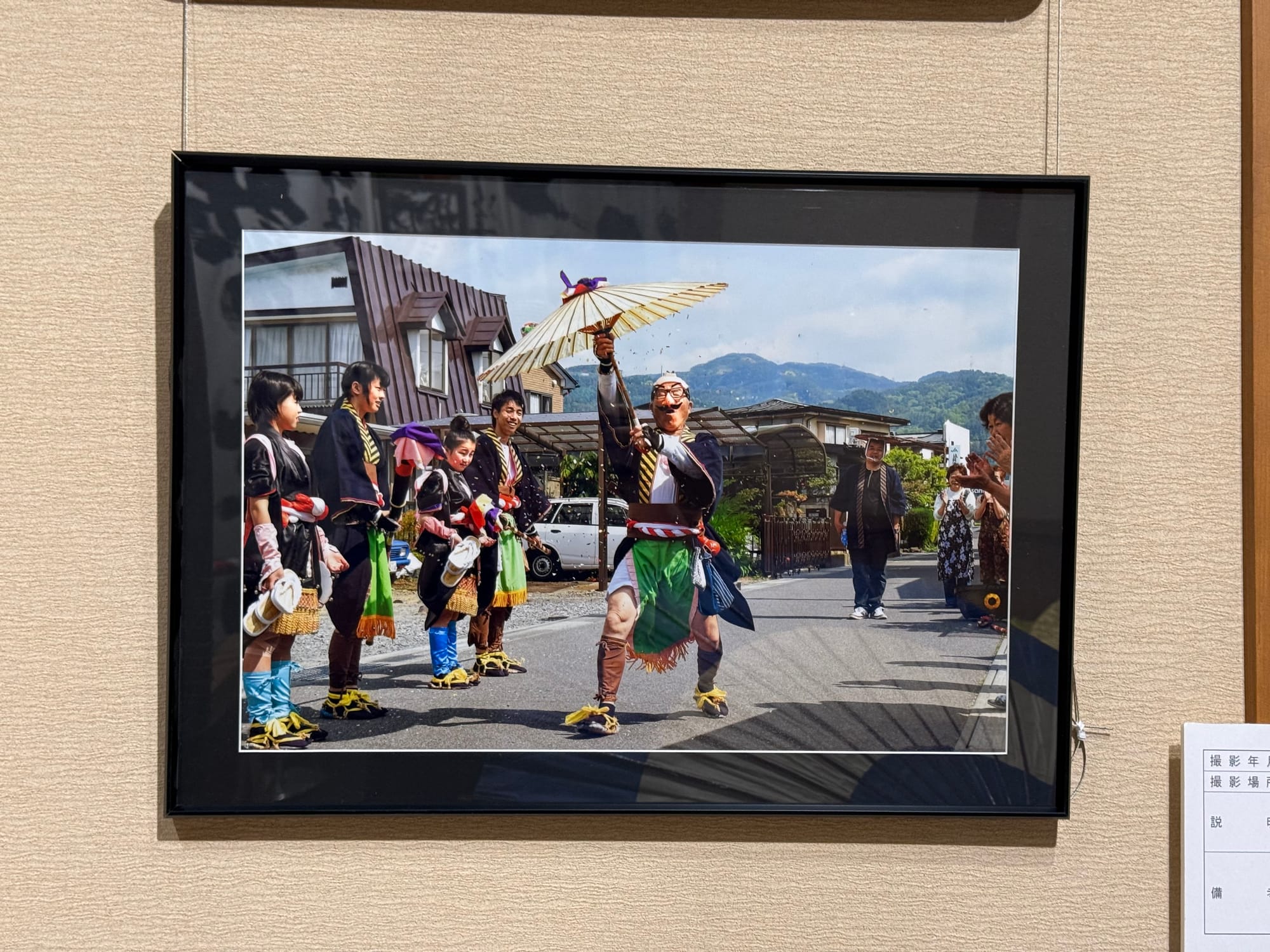 Framed photograph at Onbashira-kan Yoisa depicting a traditional festival performer dancing with a paper umbrella, surrounded by participants in costume during a local celebration.