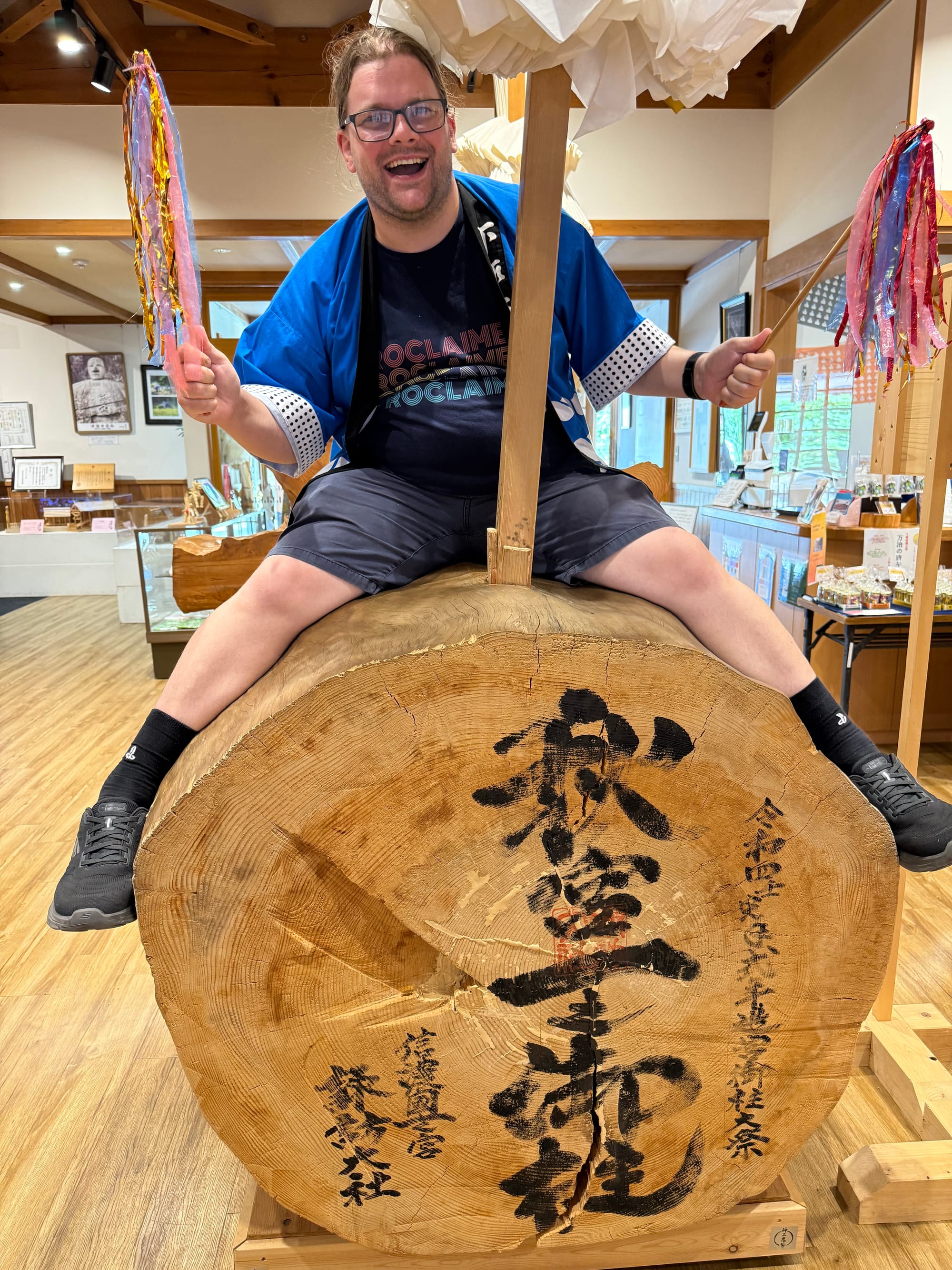 Visitor sitting on a full-sized Onbashira log at Onbashira-kan Yoisa, holding festival decorations and demonstrating the position taken by participants during the Onbashira Festival.