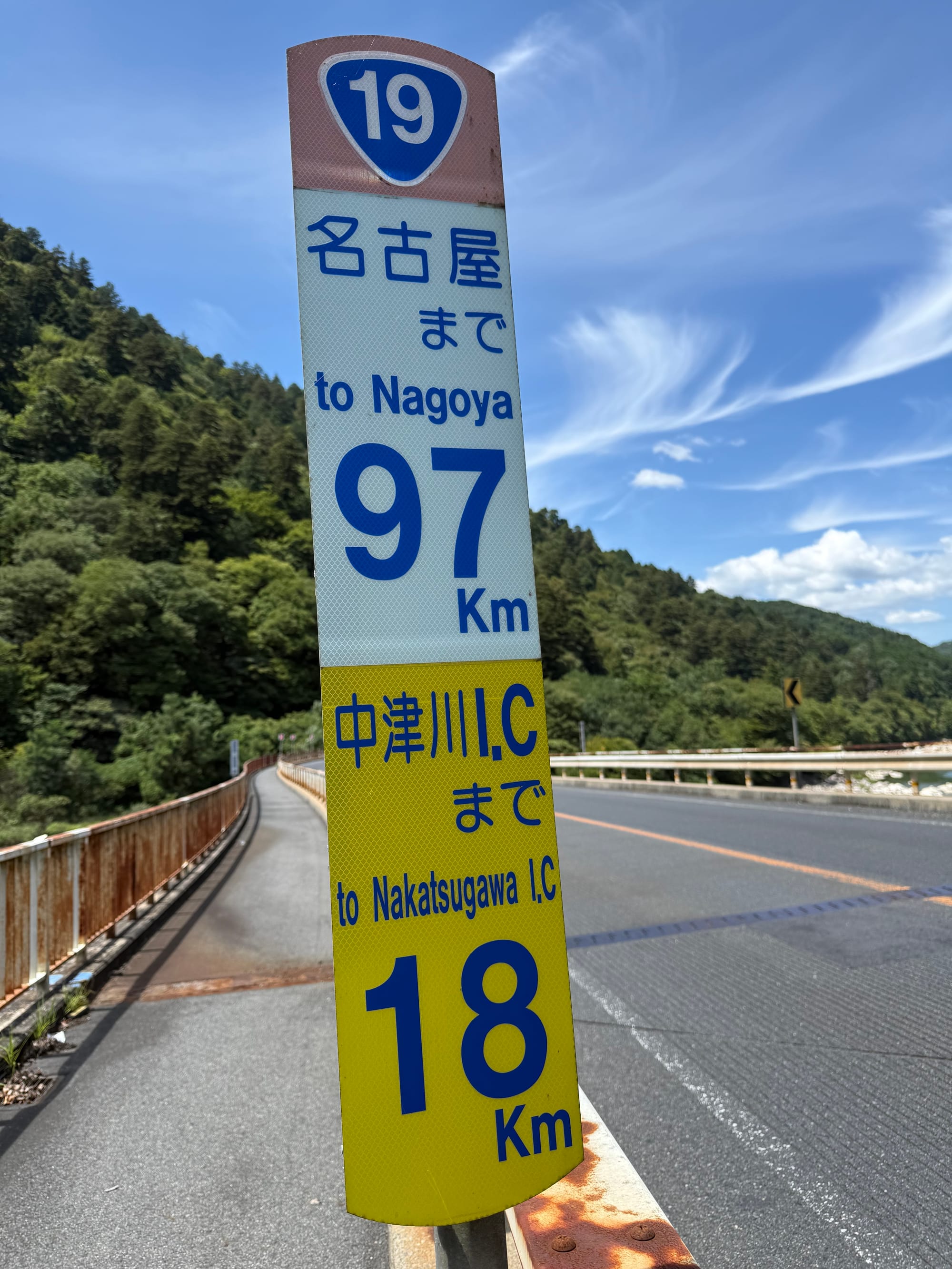 Route 19 distance marker showing 97 kilometres to Nagoya and 18 kilometres to Nakatsugawa Interchange, beside a mountain highway under a bright blue summer sky.