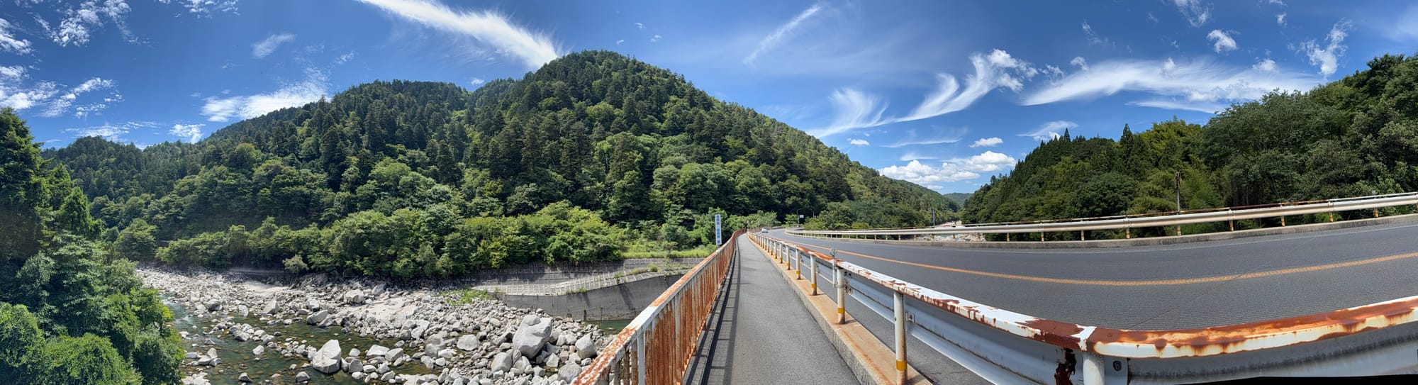 National Route 19 crossing a rocky river valley in the Kiso region, with forested mountains, a clear mountain stream and wispy clouds stretching across a bright blue sky.
