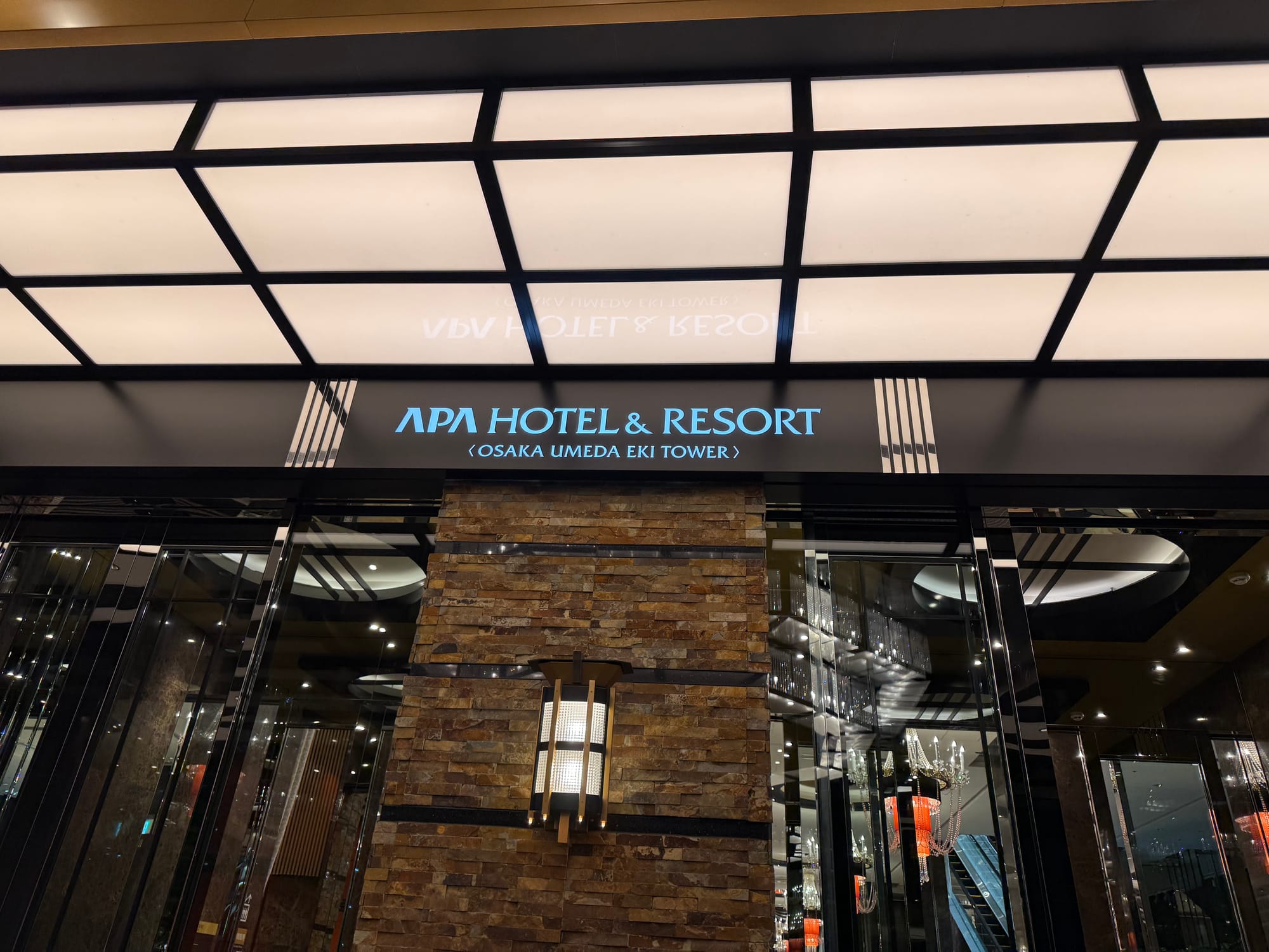 Entrance of APA Hotel & Resort Osaka Umeda Eki Tower at night, featuring illuminated signage, glass doors, stone cladding and modern architectural details.