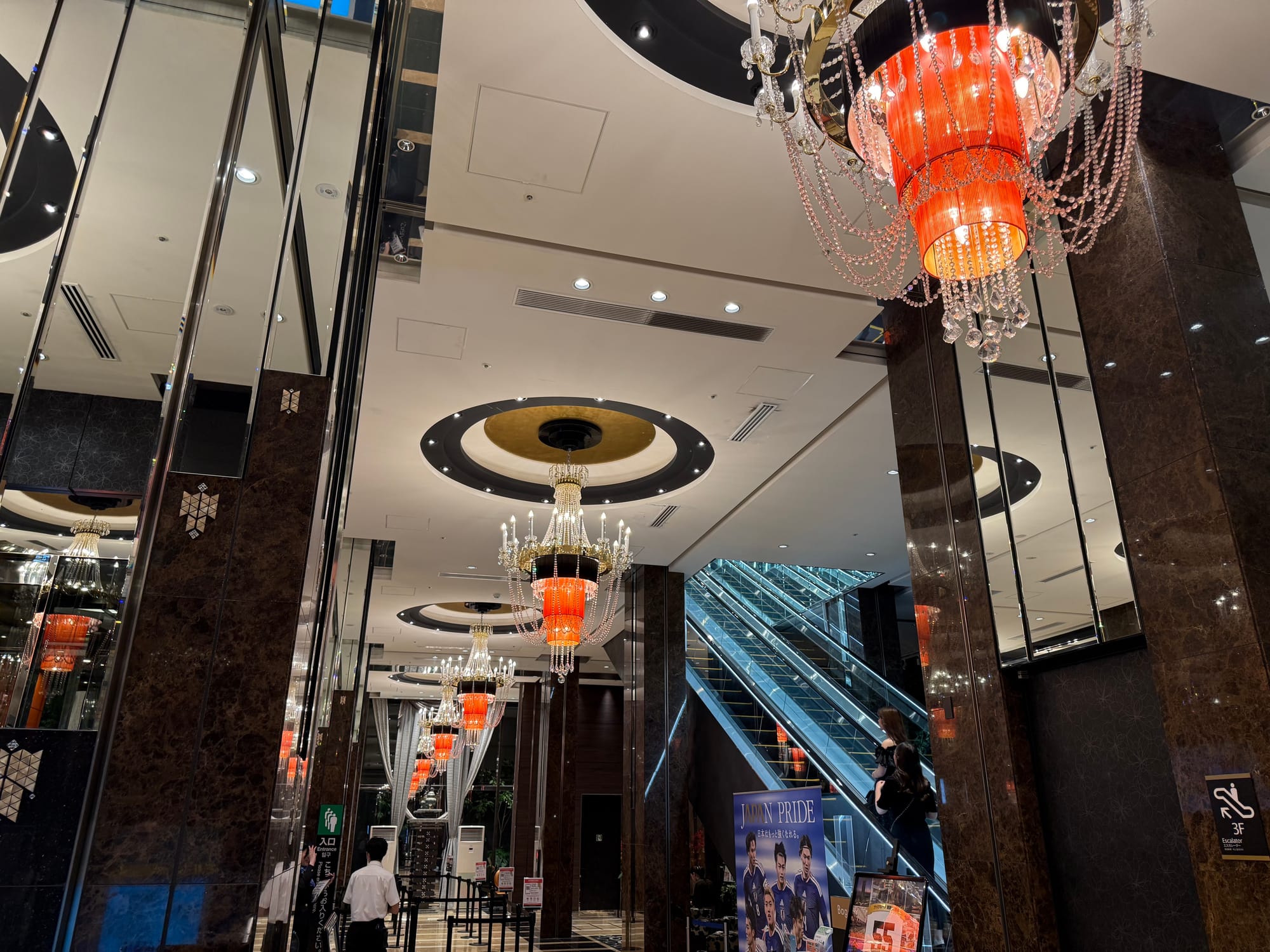 Grand lobby of APA Hotel & Resort Osaka Umeda Eki Tower, featuring red chandeliers, mirrored columns, escalators and an ornate modern interior design.