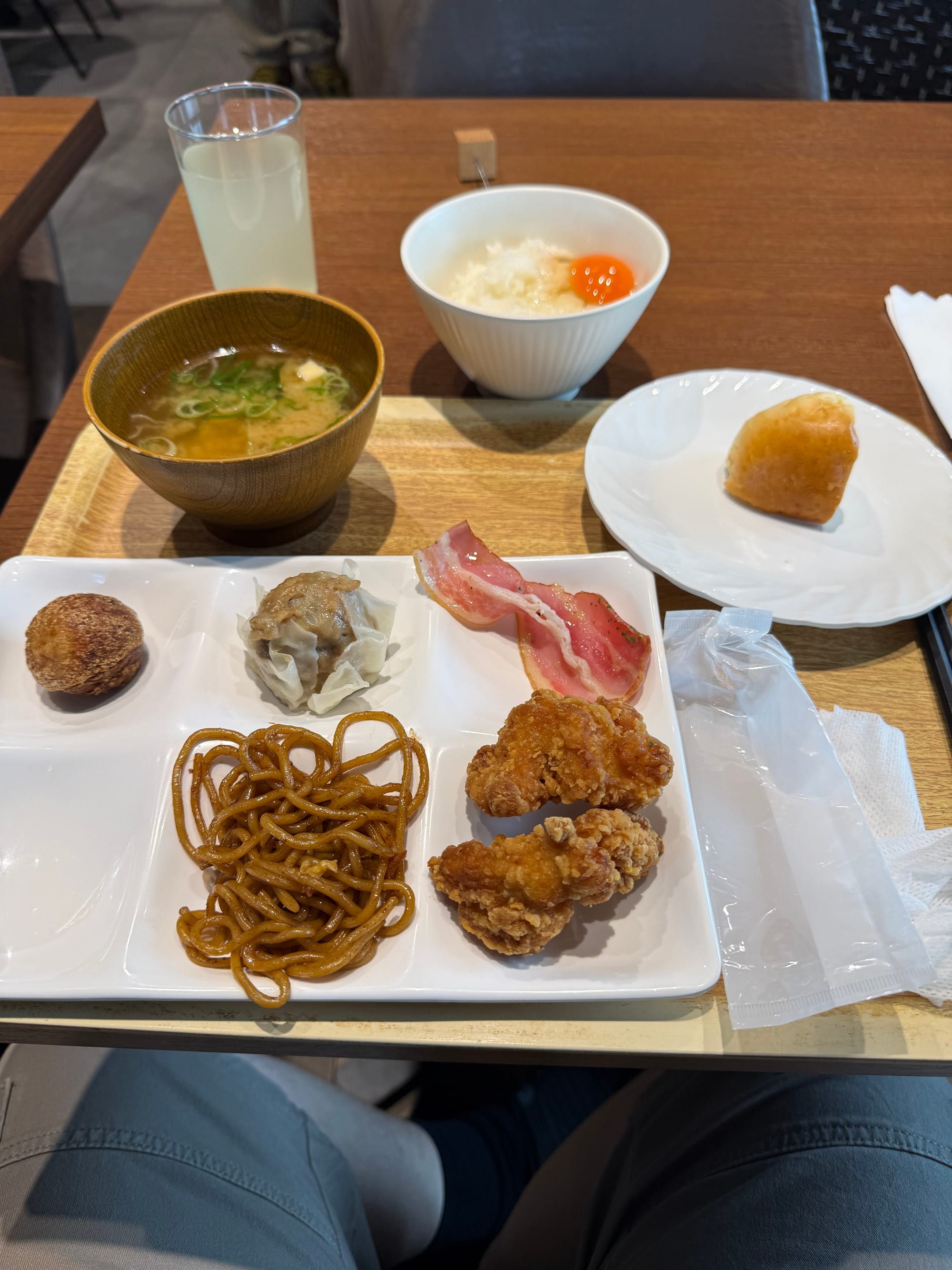 Hotel breakfast in Osaka featuring rice topped with a raw egg, miso soup, karaage chicken, bacon, shumai, fried noodles, bread roll and a glass of juice.