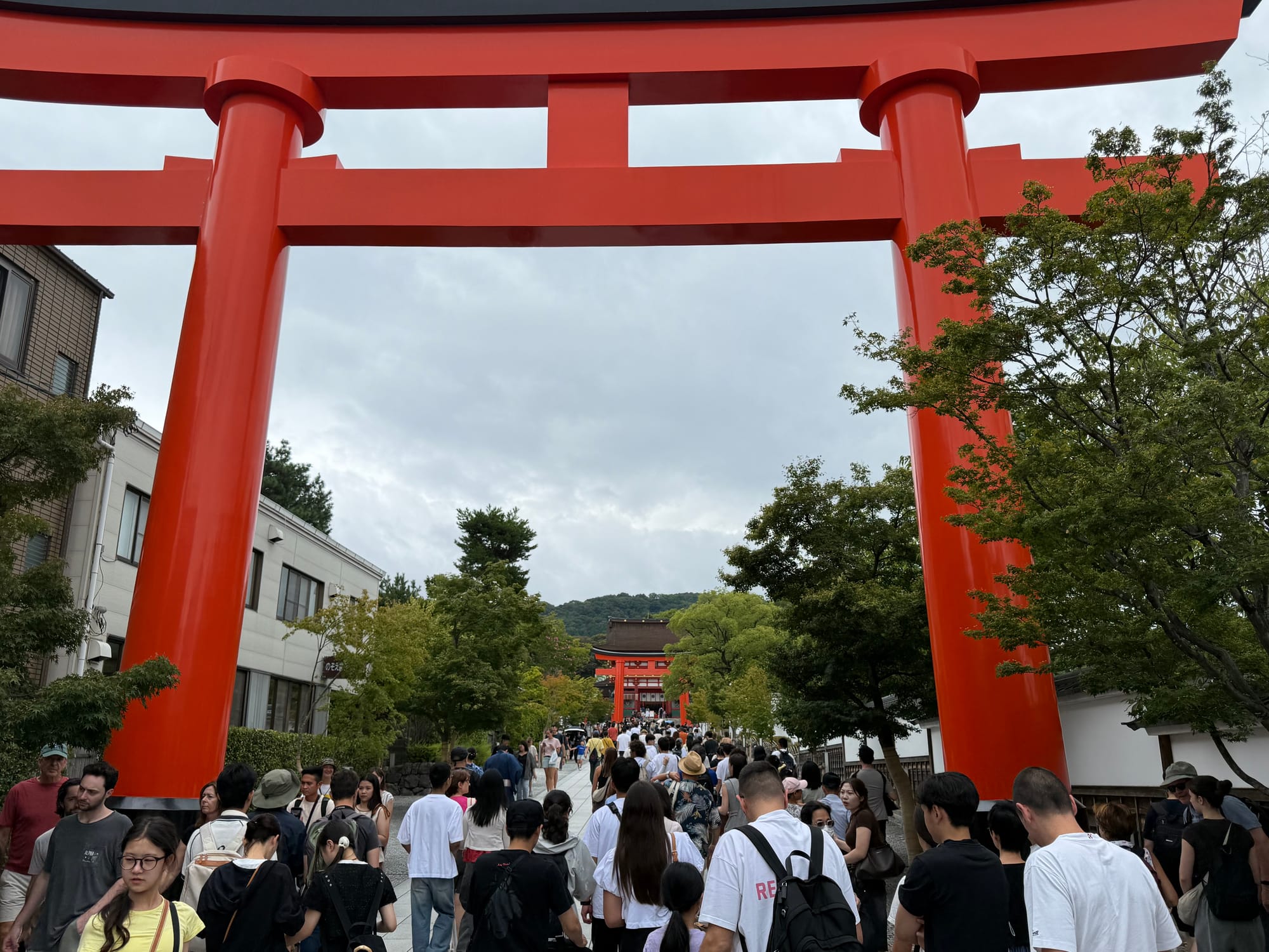 Visitors walk through the giant vermilion torii gate at Fushimi Inari Taisha in Kyoto, with crowds gathering along the shrine approach beneath overcast skies.