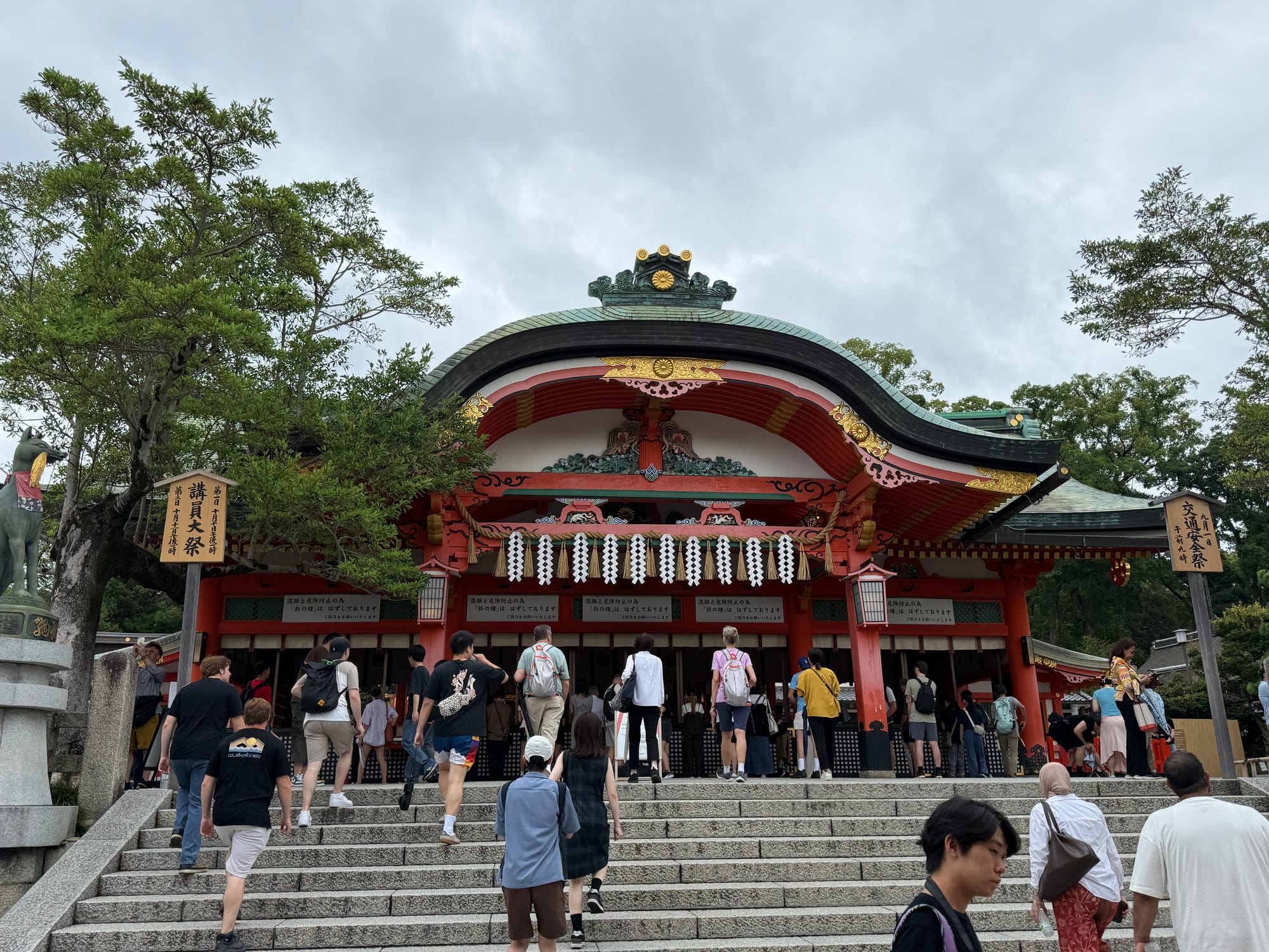 Visitors climb the steps to the main worship hall of Fushimi Inari Taisha in Kyoto, with the shrine's ornate vermilion architecture standing beneath dark, overcast skies.
