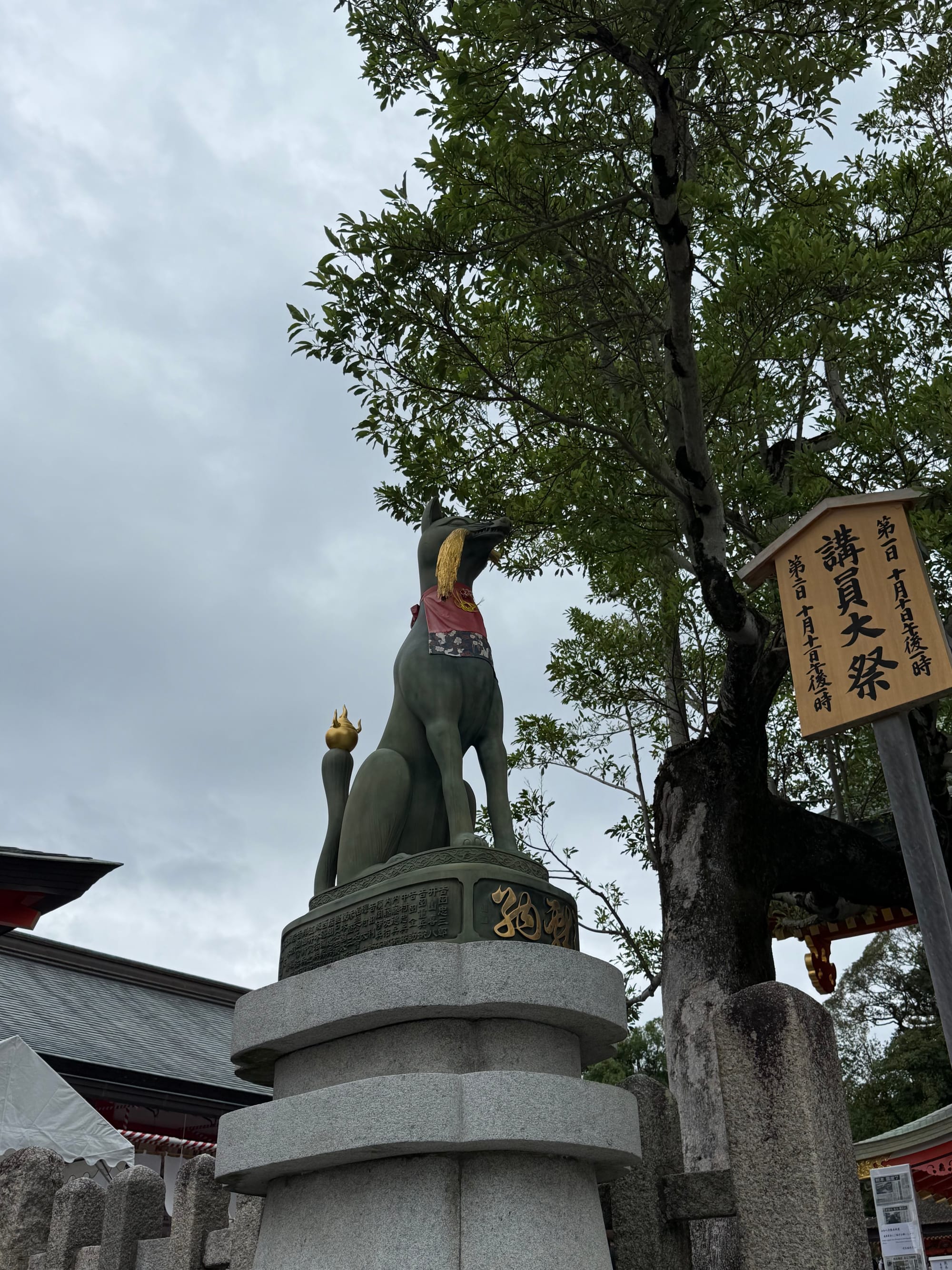 Bronze fox statue at Fushimi Inari Taisha in Kyoto, standing on a stone pedestal and wearing a red bib, with shrine buildings, trees and festival signage nearby.