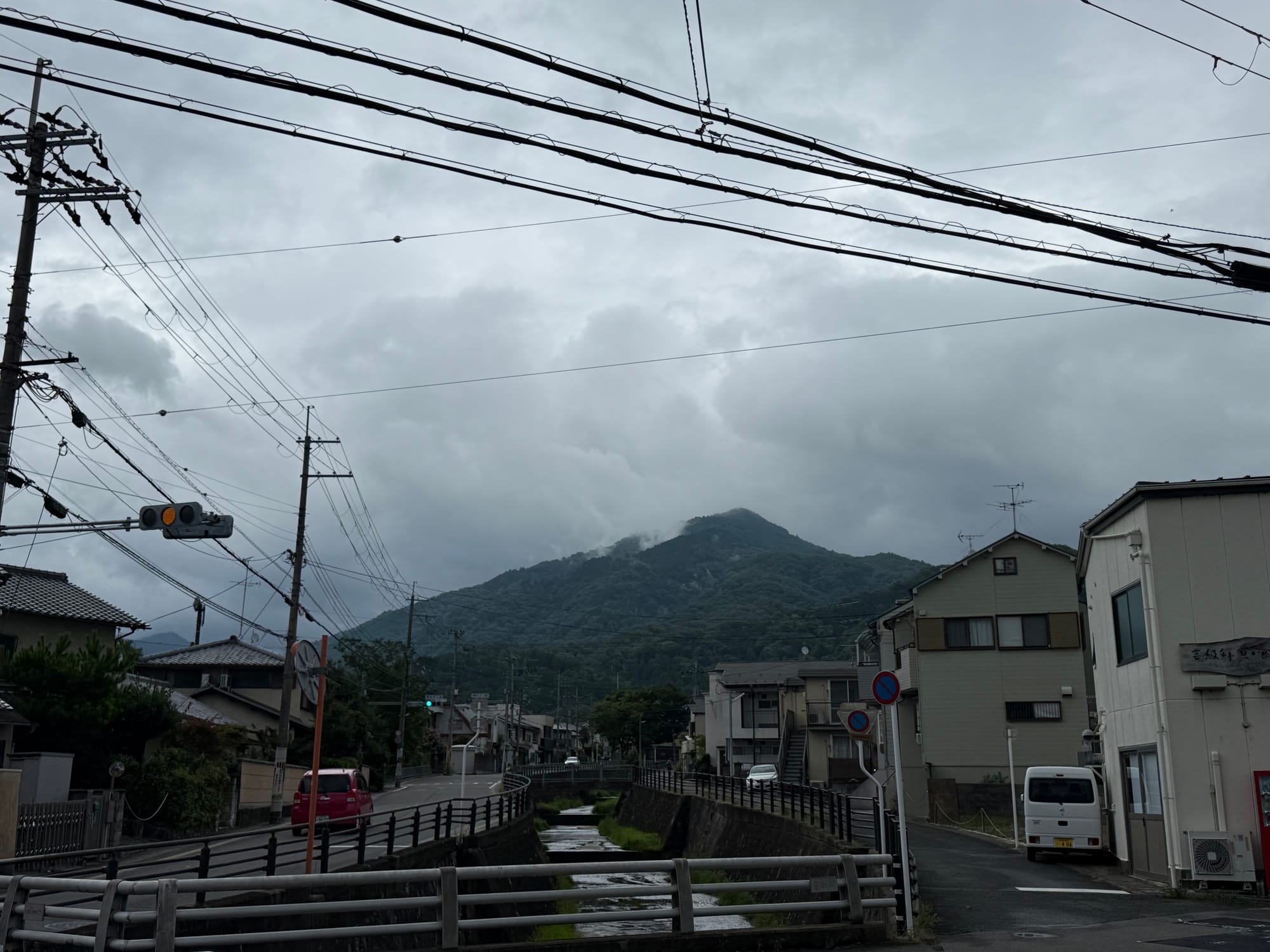 View toward Mount Hiei from southern Kyoto beneath low rain clouds, with a canal, residential streets, power lines and forested mountain slopes under grey skies.