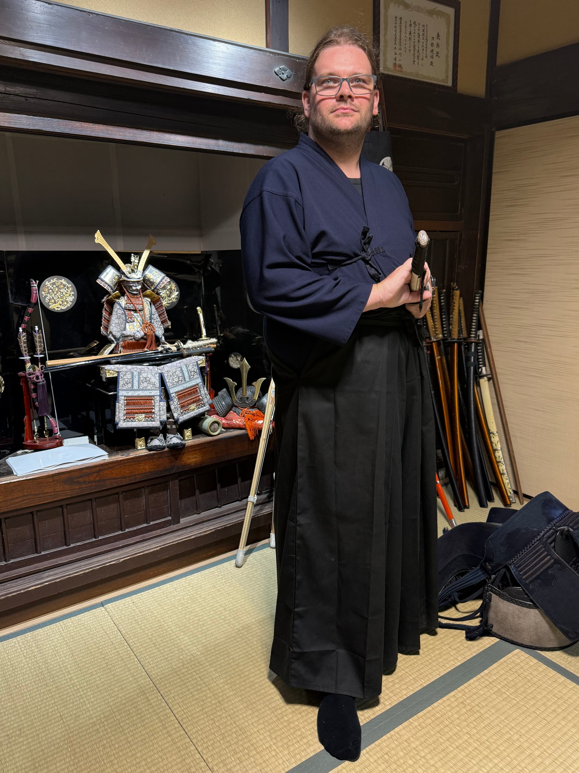 Visitor dressed in traditional martial arts attire holding a sheathed katana in a tatami room in Kyoto, with samurai armour, practice weapons and kendo equipment on display.