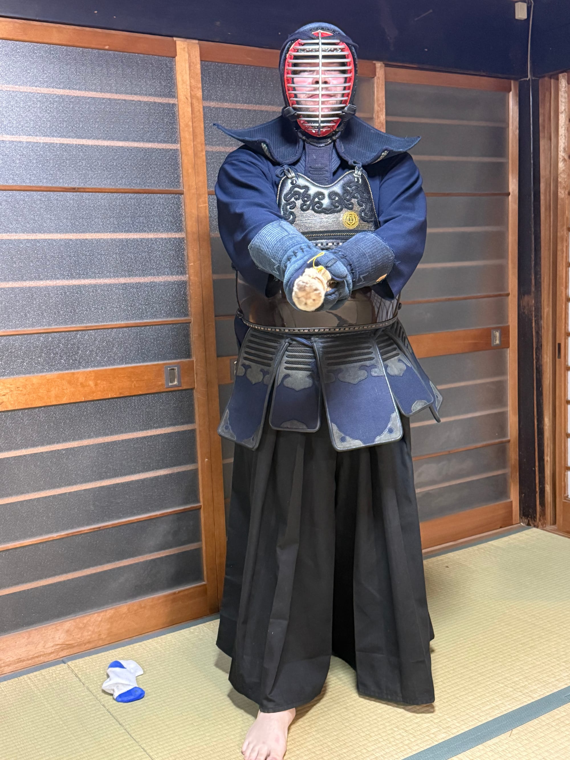 Visitor wearing full kendo armour in a traditional Kyoto dojo, holding a bamboo shinai in a ready stance, with tatami mats and sliding shoji screens in the background.